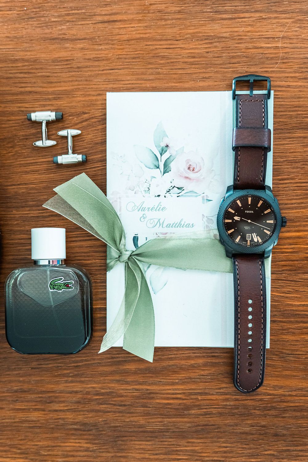 Wedding flat lay featuring a Lacoste cologne, cufflinks, floral invitation, green ribbon, and brown leather watch.