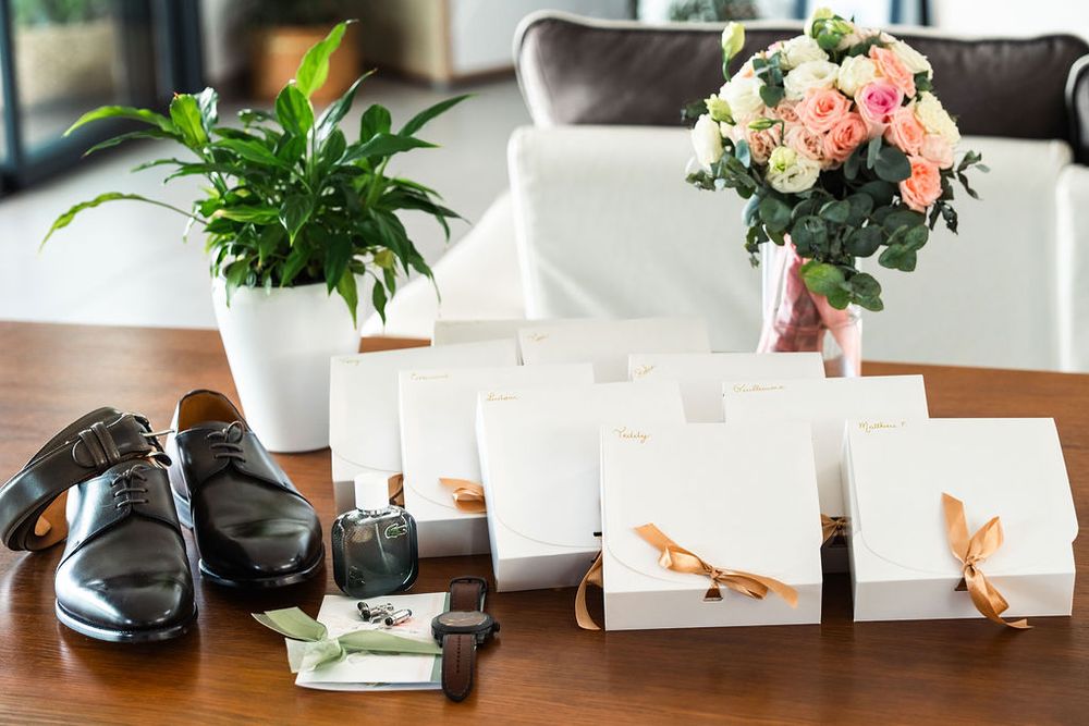 Elegant wedding flat lay with black dress shoes, gift boxes with ribbon, floral bouquet, and accessories on a wooden table.