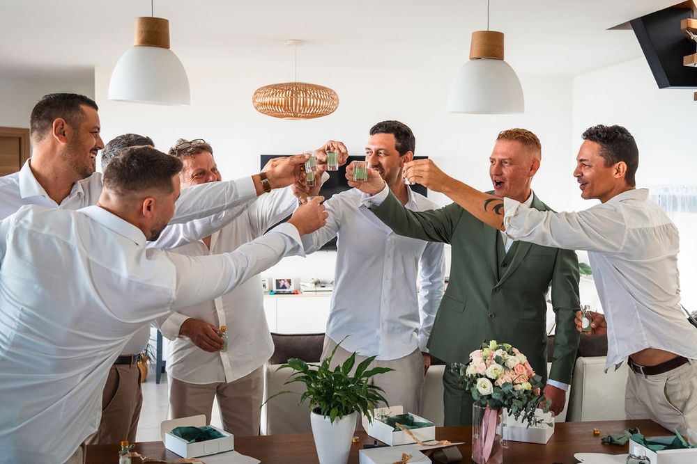 Group of groomsmen in white shirts toasting drinks with groom in green suit at wedding celebration.