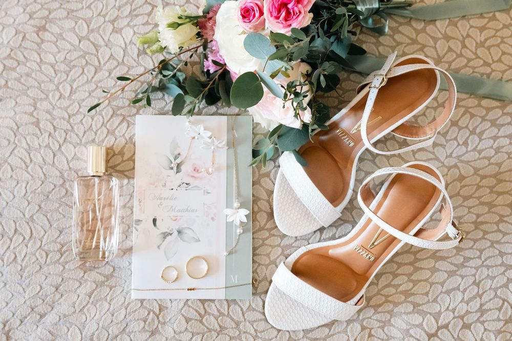 White bridal heels, wedding rings, floral bouquet, and elegant invitation suite arranged on a textured surface.