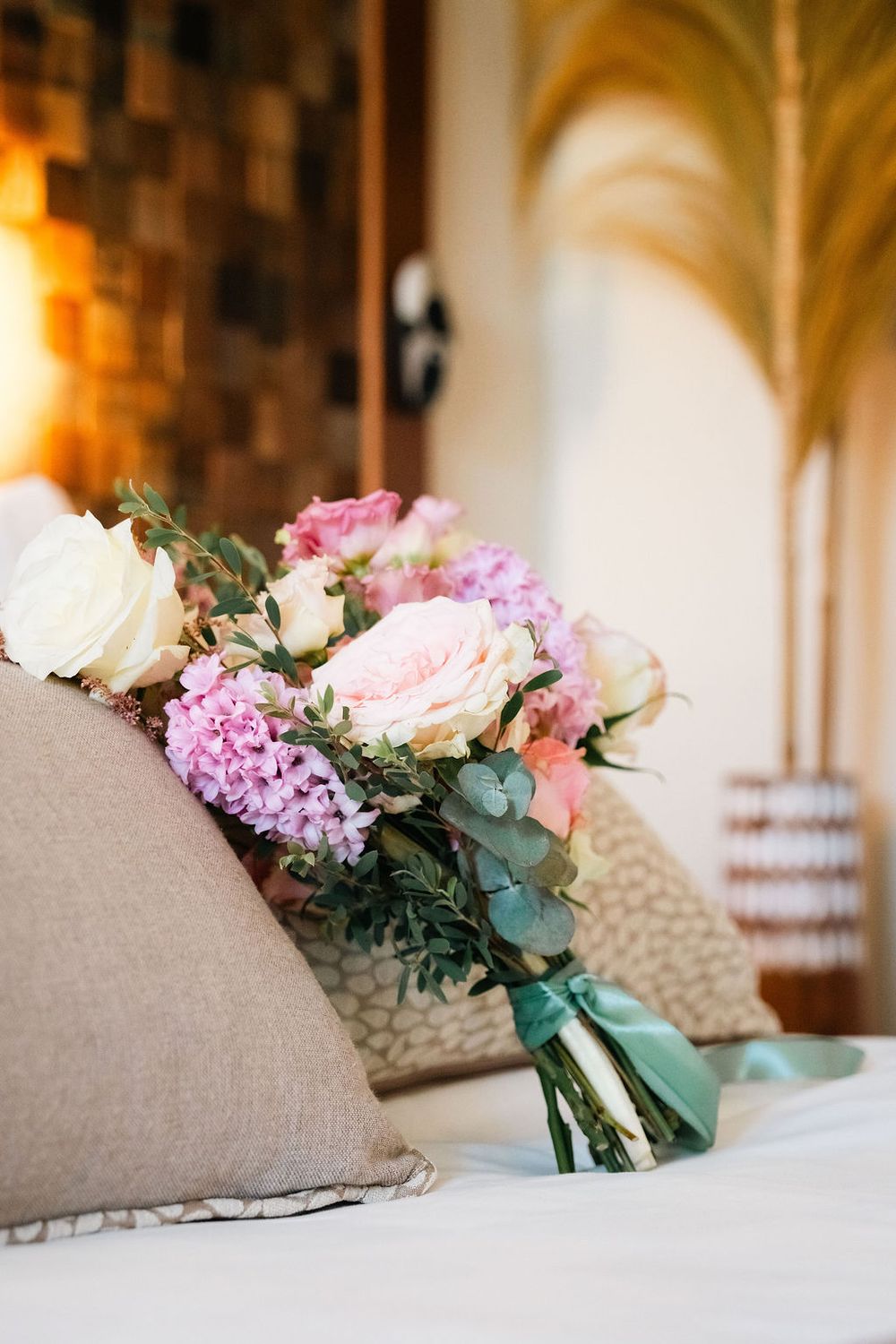 Romantic bridal bouquet of pink roses, hydrangeas, and eucalyptus resting against beige pillows on a white bed.