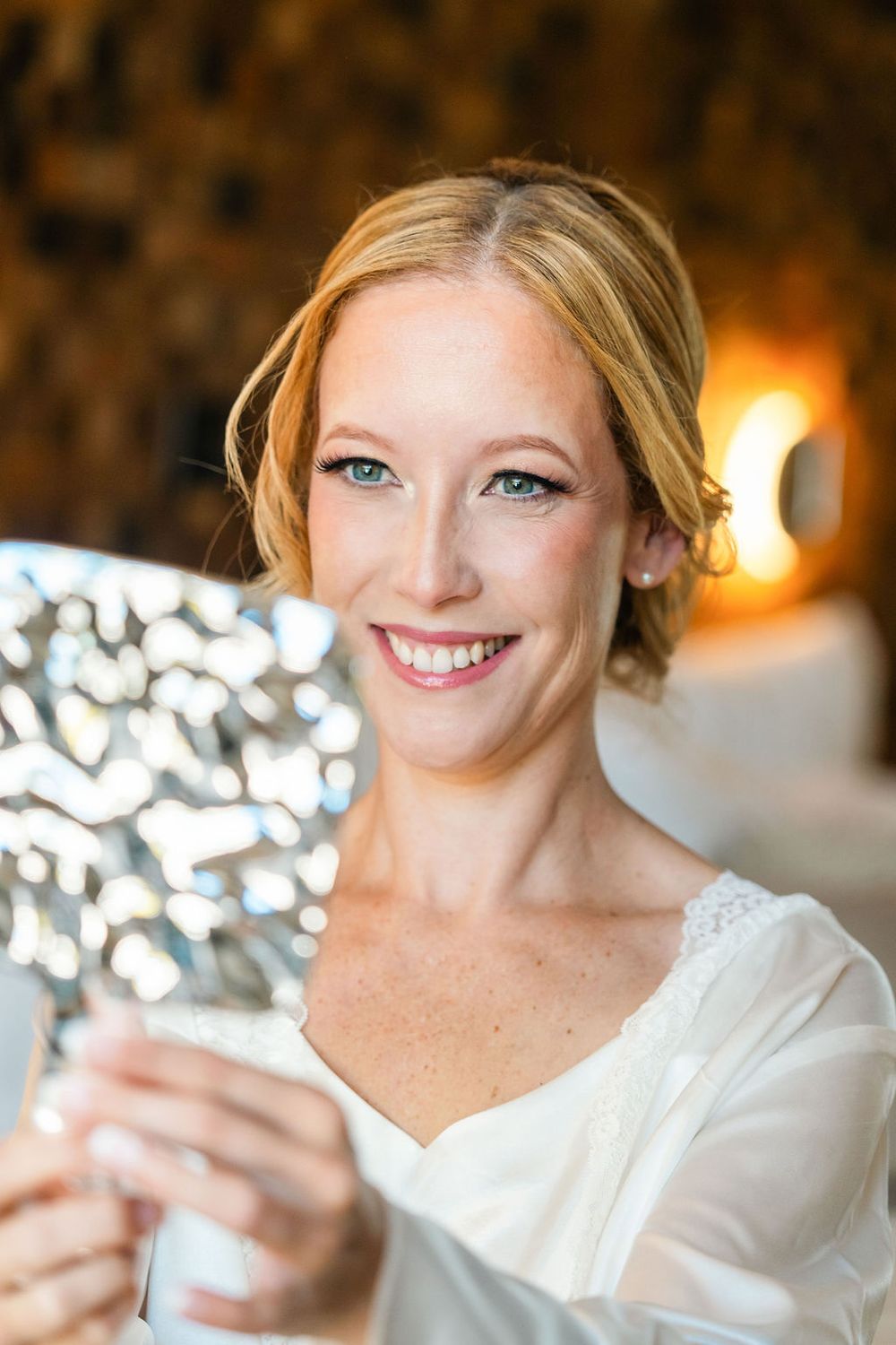 Smiling blonde woman with blue eyes holding a decorative silver mirror, wearing a white top in a warmly lit room.