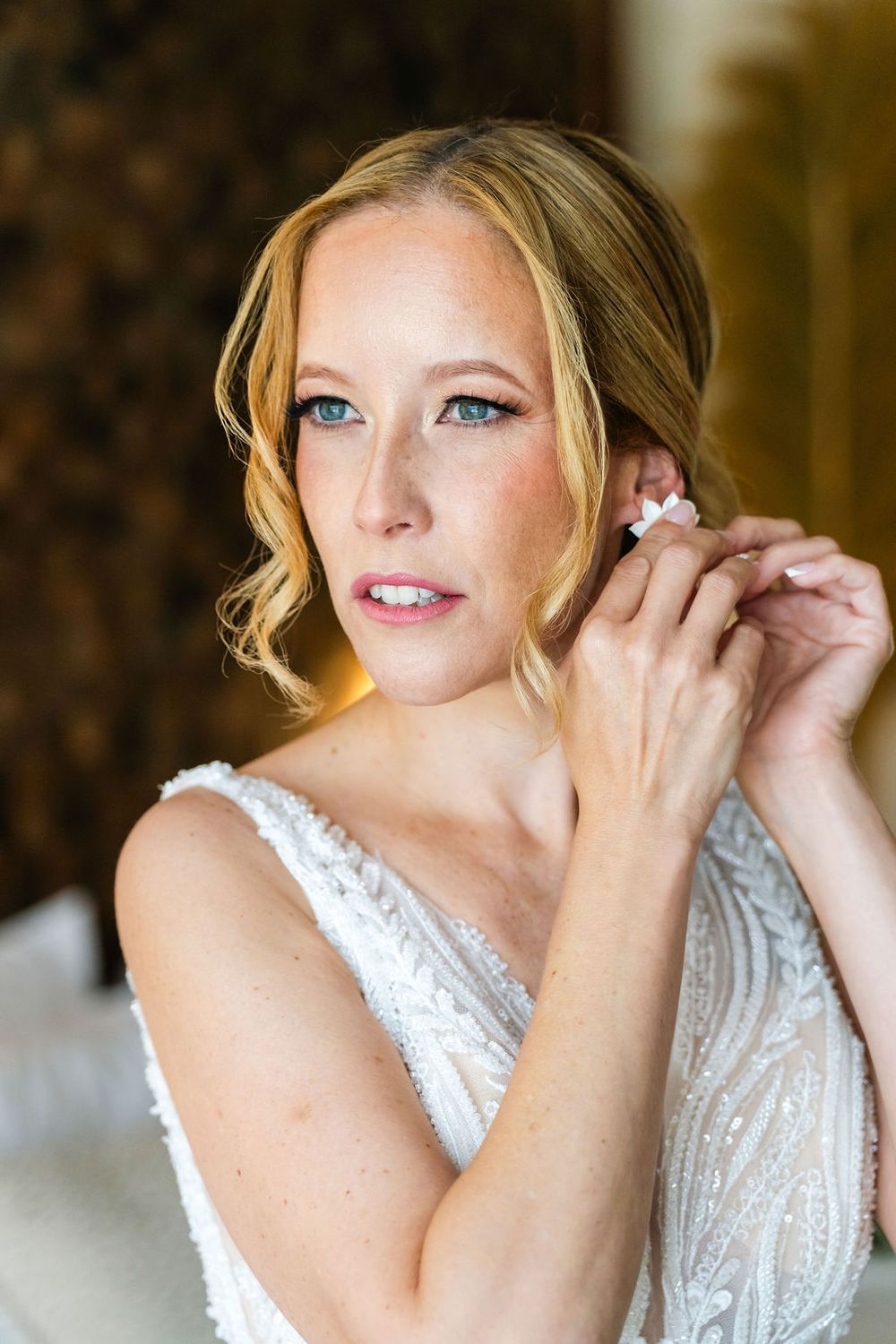 Blonde bride with blue eyes putting on earrings in a white beaded wedding gown, elegant bridal portrait.