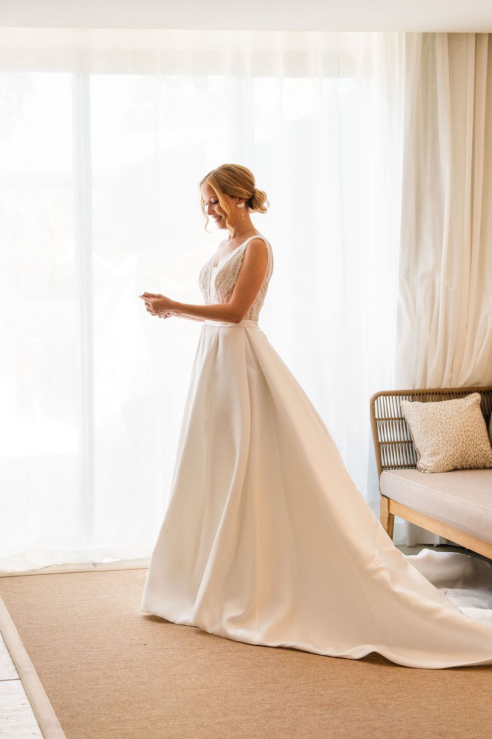 Bride in elegant white ball gown wedding dress standing by sheer curtained window in bright room.