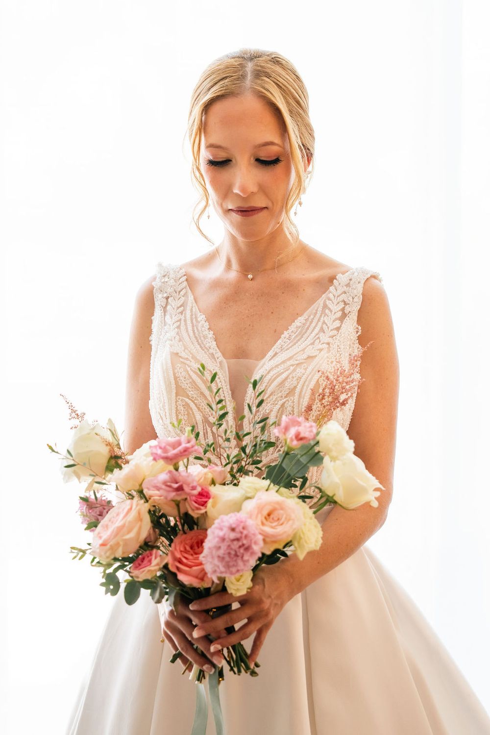 Elegant bride in a white lace gown holding a pastel pink and cream rose bouquet, looking down gracefully.