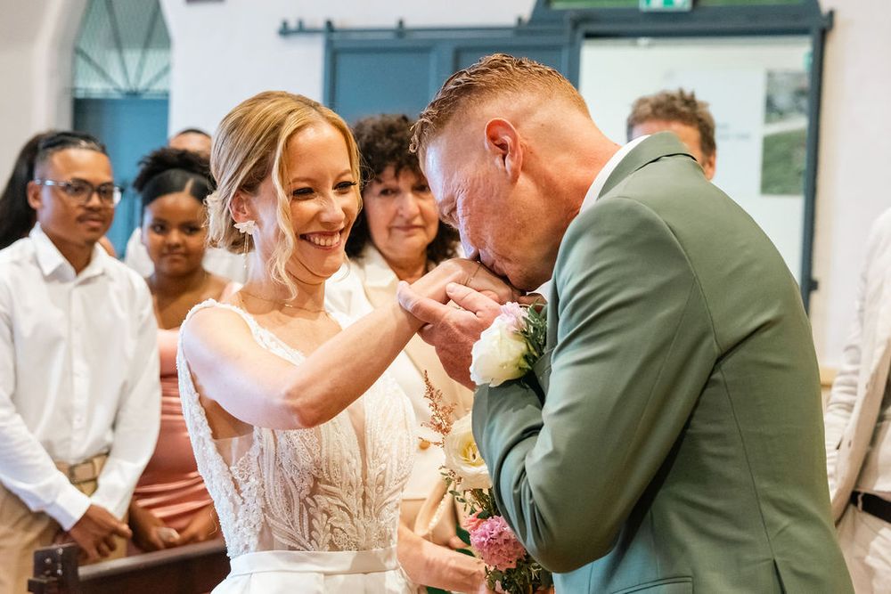 Groom in sage green suit kisses bride's hand during emotional wedding ceremony with guests watching in background.