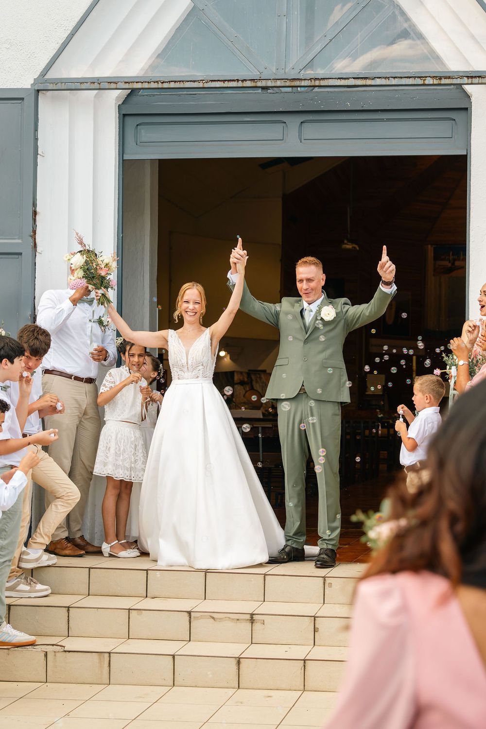Bride and groom exit church with raised hands, surrounded by cheering guests blowing bubbles on sunny wedding day.