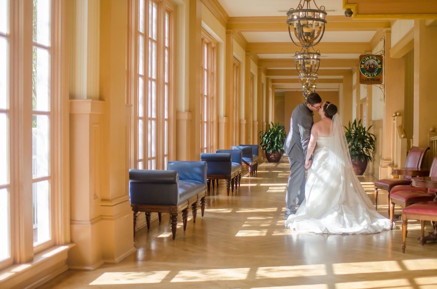 Bride and groom share a kiss in a sunlit elegant hotel corridor with blue benches and tall windows on their wedding day.