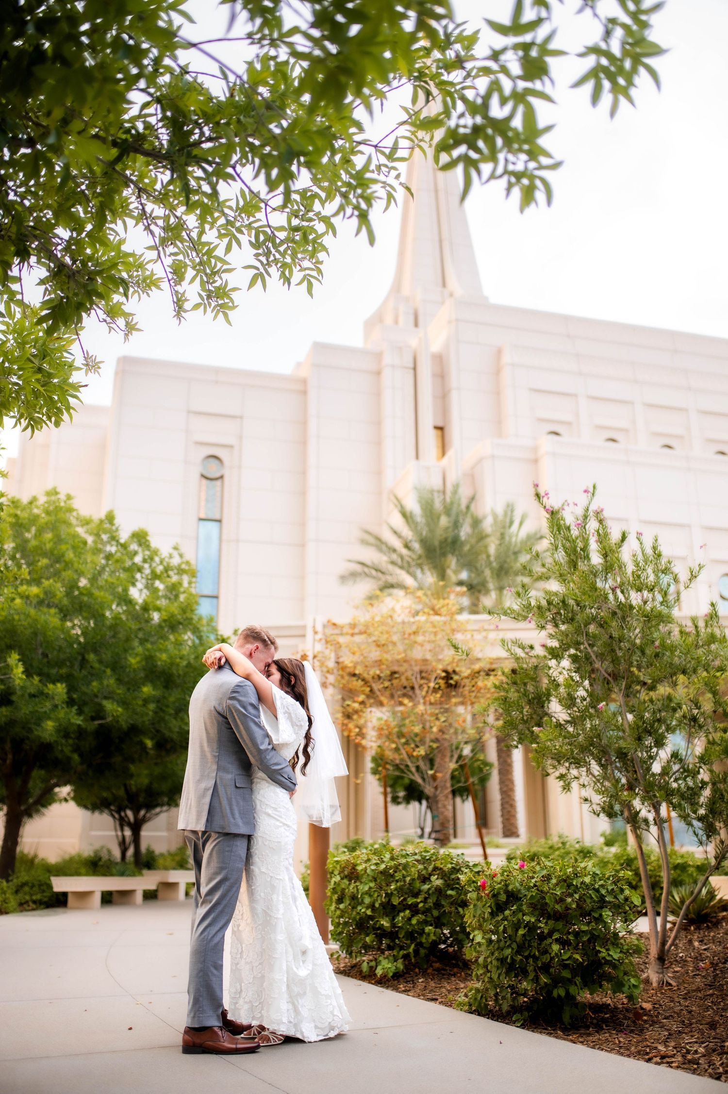 Bride and groom share a tender embrace on a garden path with an LDS temple behind them.