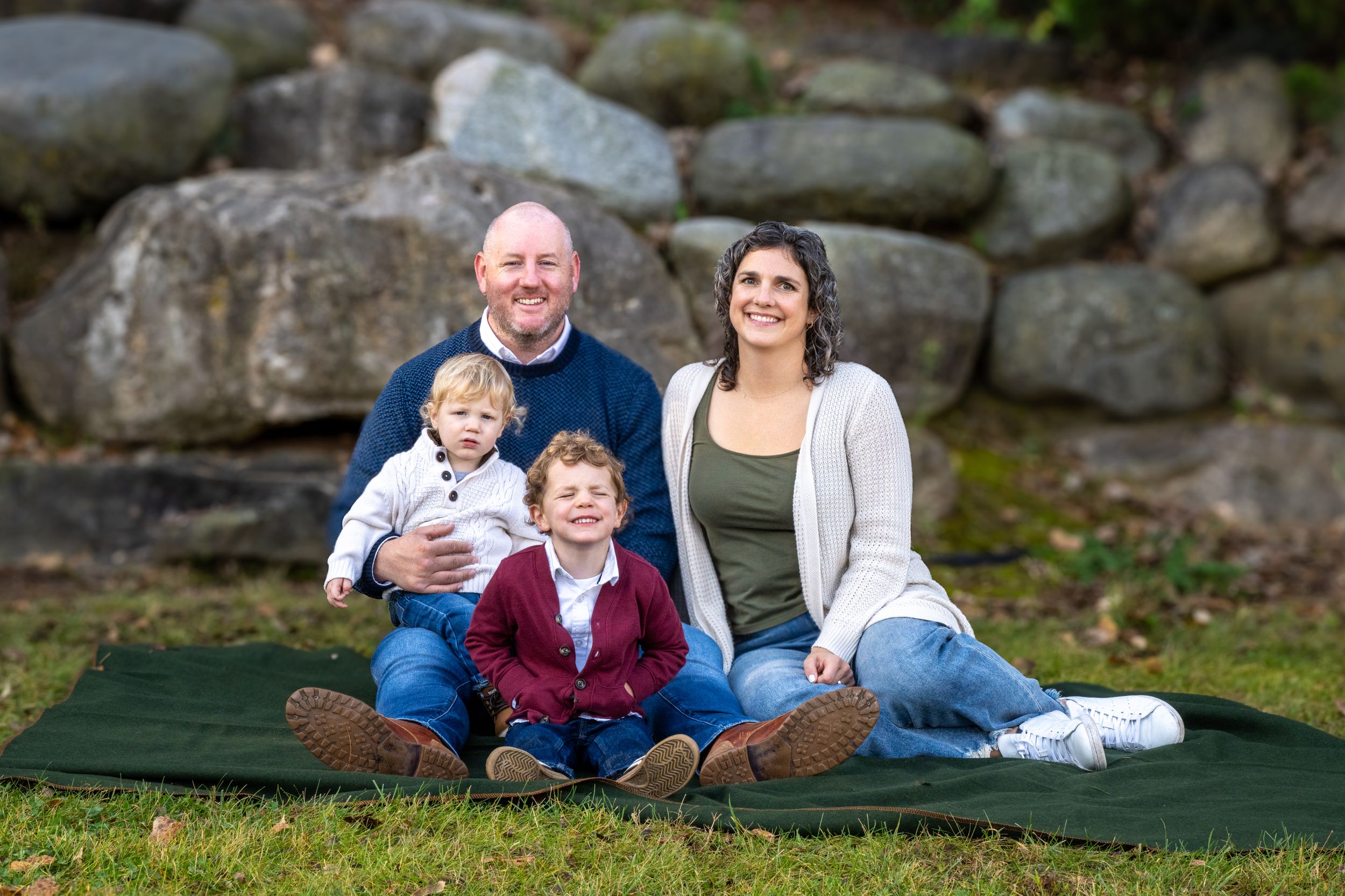 Family of four smiling outdoors on green blanket with stone wall backdrop during fall season portrait session.