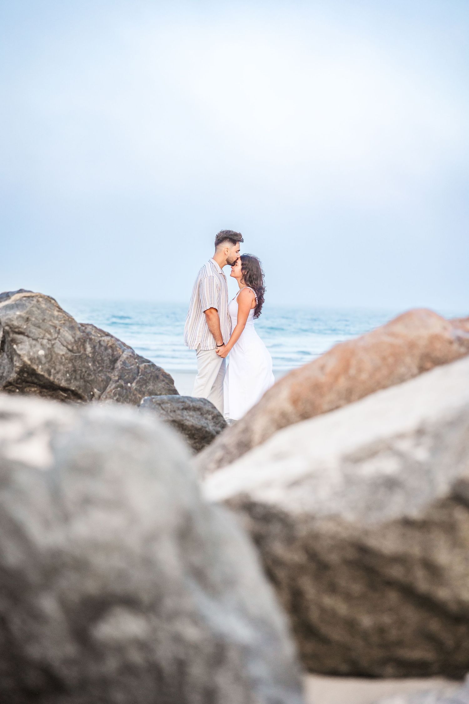 Romantic couple embracing on a rocky beach with ocean waves in the background, dressed in white outfits.
