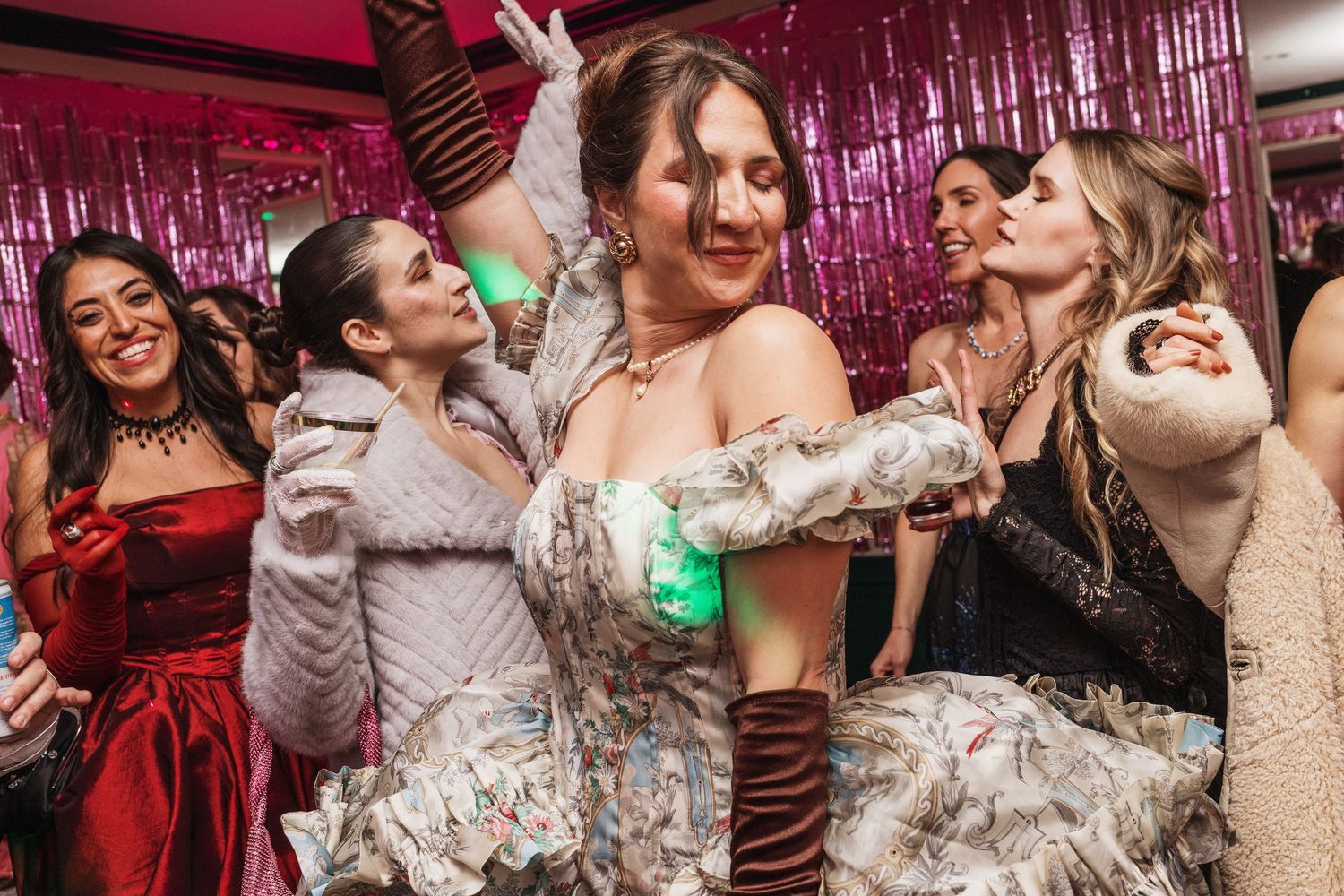 Group of women dancing and celebrating at a glamorous party with pink tinsel backdrop decorations.