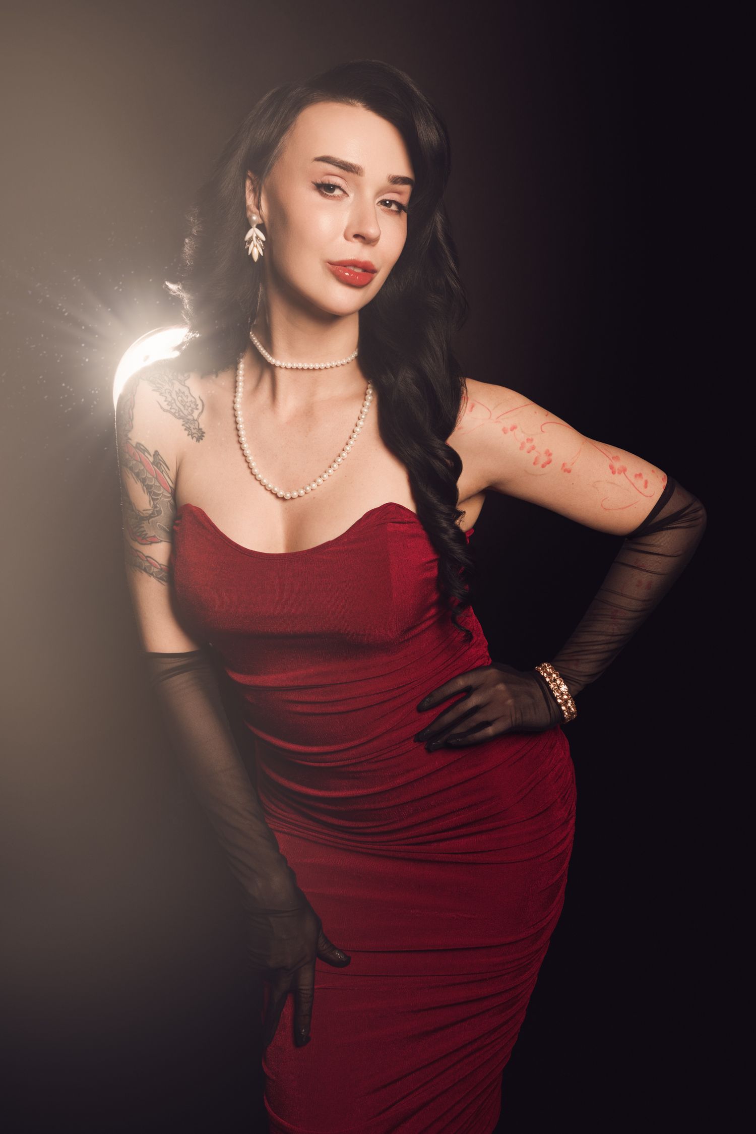 Elegant woman in red dress with pearl necklace, tattoos, and black gloves posing against dark background.