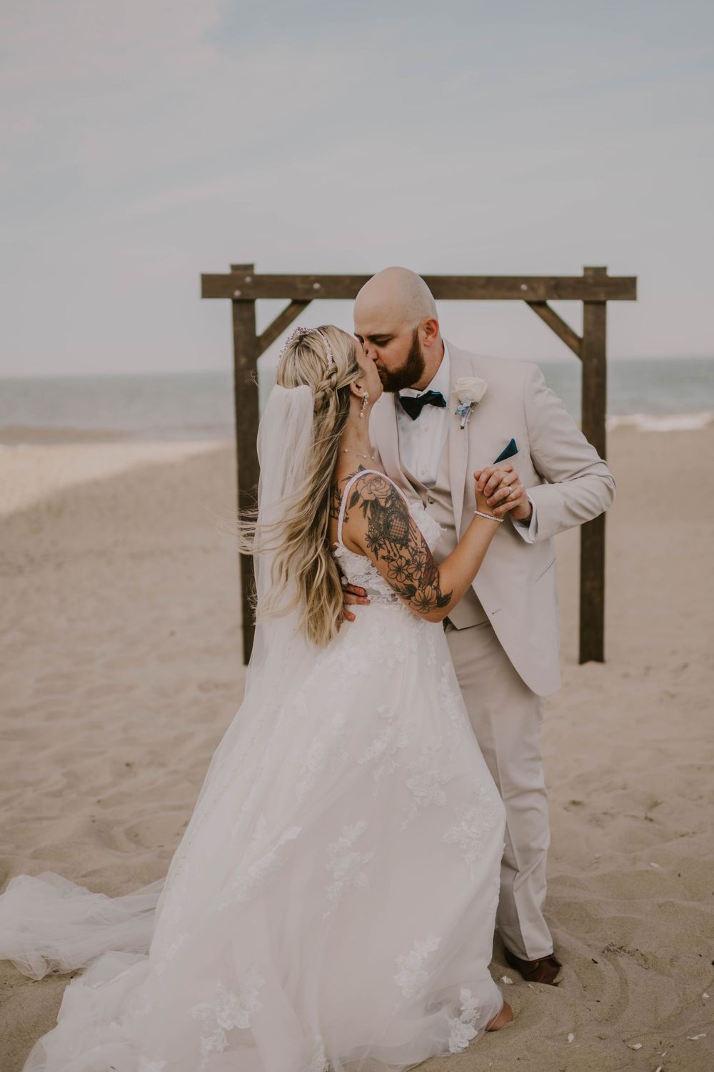Bride and groom share first kiss on sandy beach, she in white gown, he in light suit with bow tie, wooden arch behind.