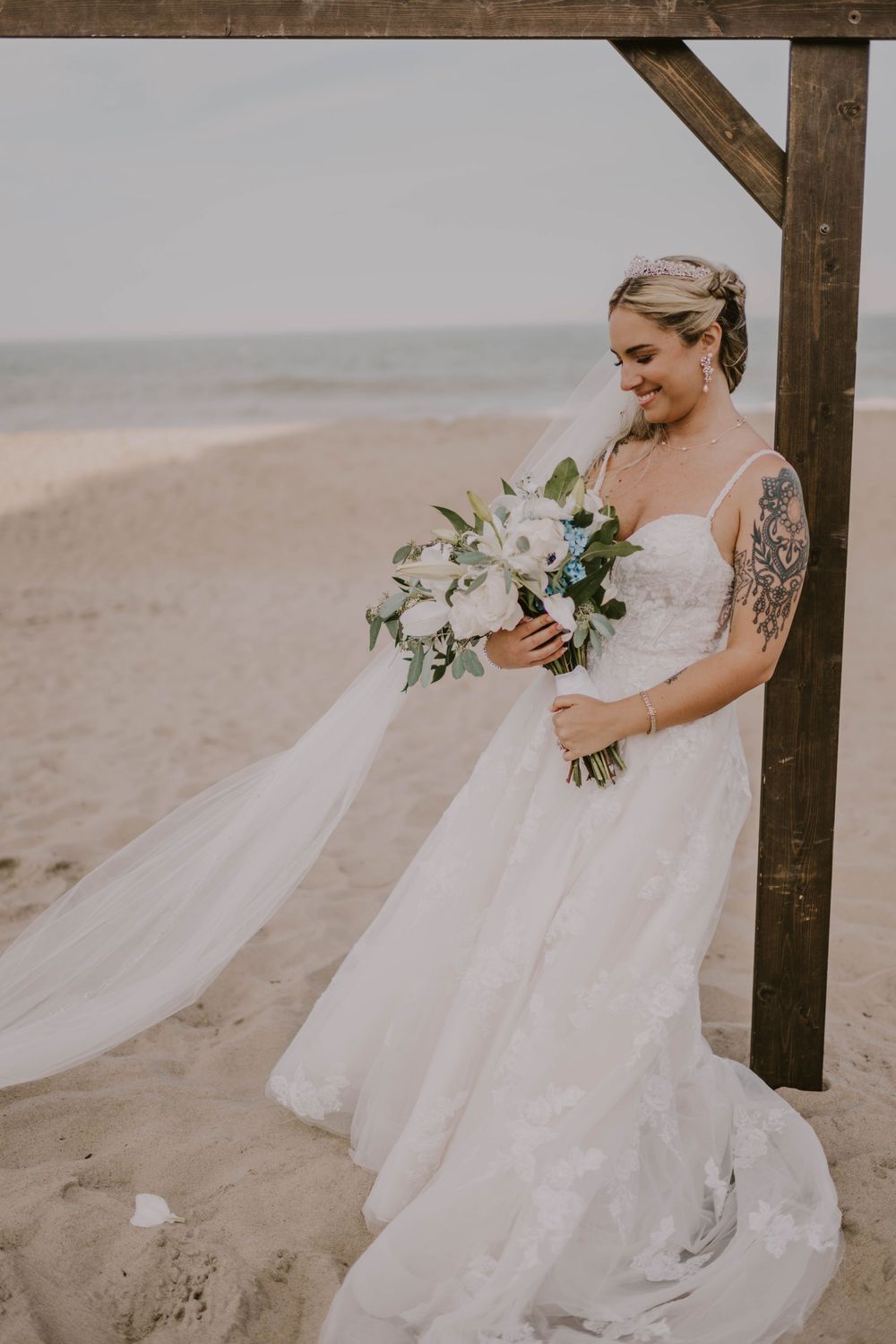 Bride in white gown holding bouquet of white and blue flowers on sandy beach near wooden arch.