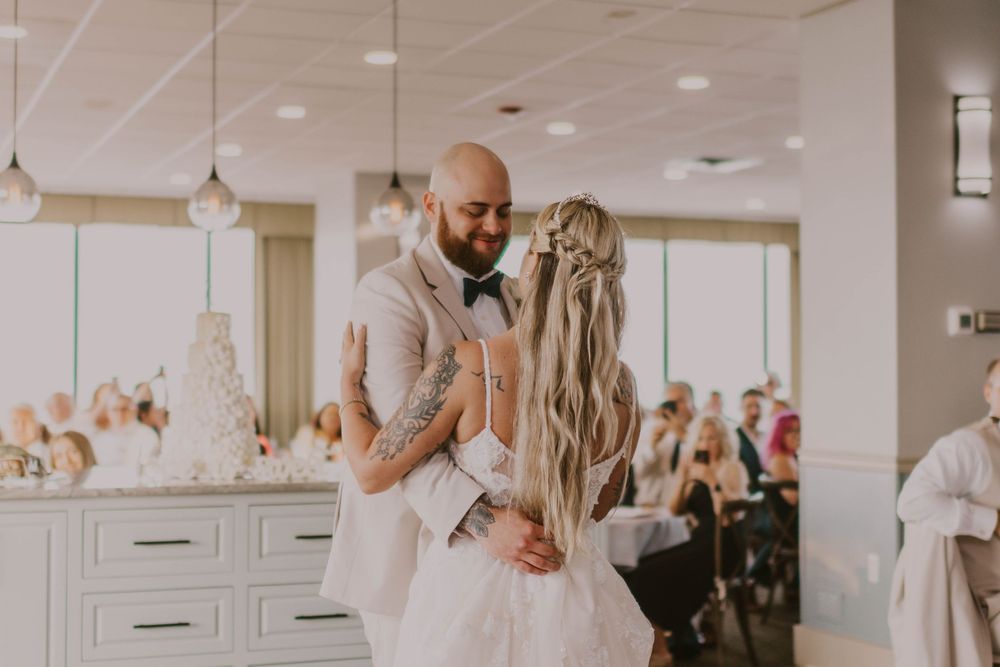 Bride and groom share first dance at wedding reception, groom with tattoos in tan suit embracing bride in white gown.