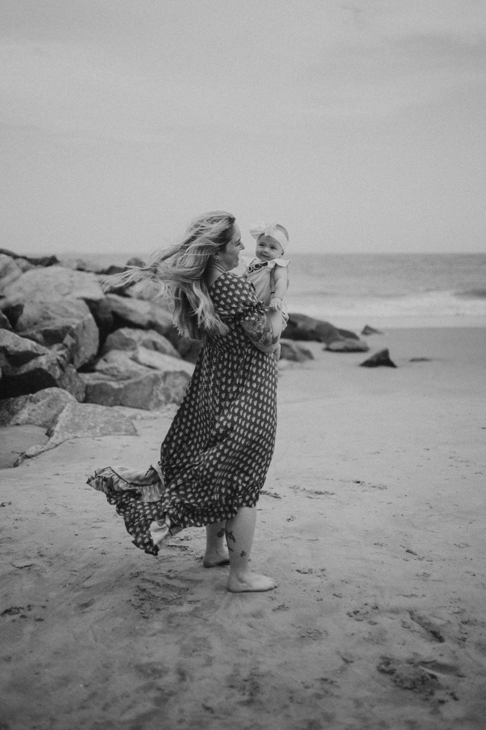 Black and white photo of a woman in a floral dress holding a baby on a sandy beach with rocks in the background.