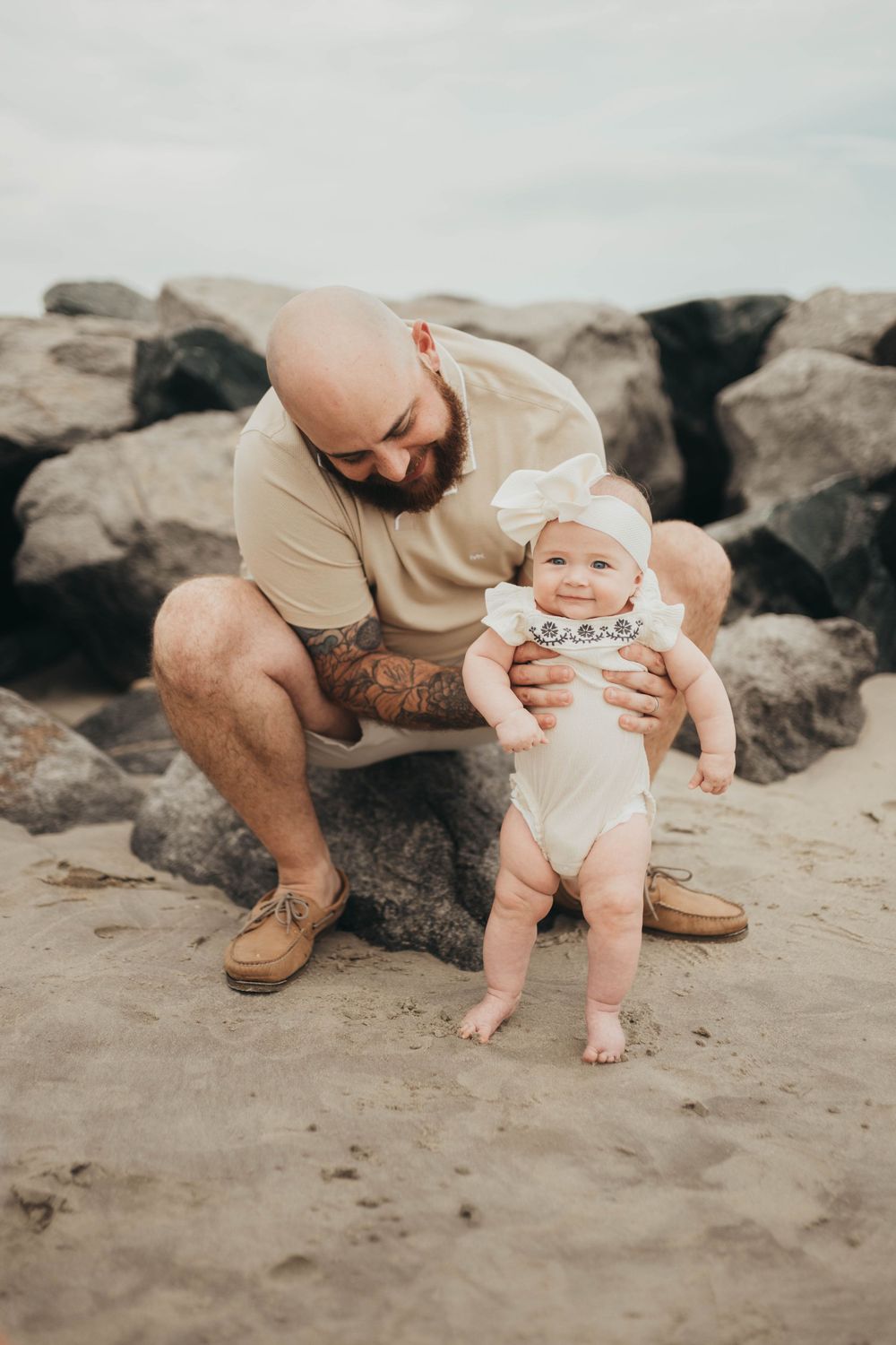 Bald tattooed father helping baby girl stand on sandy beach, rocks in background.