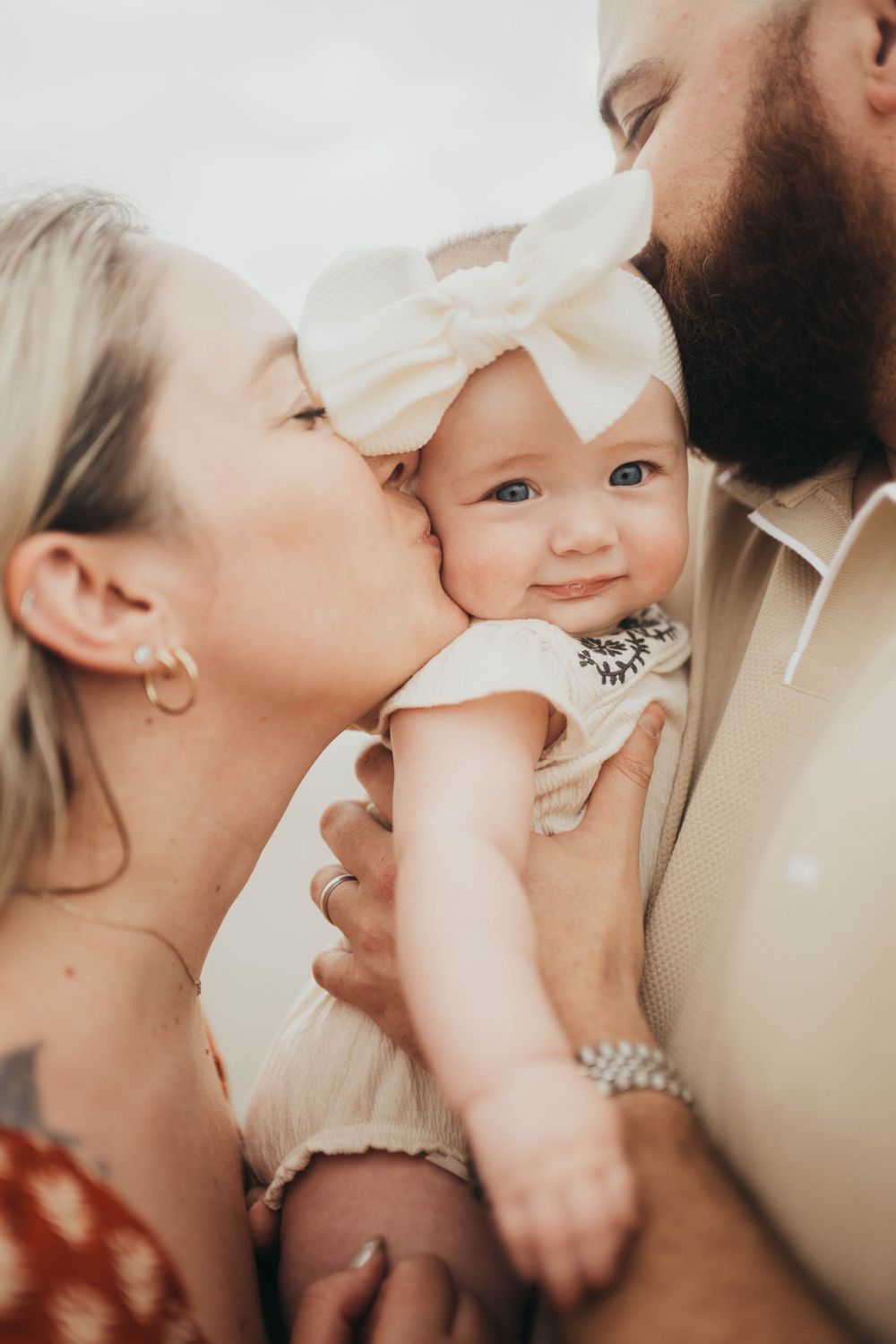 Parents kissing their smiling baby girl wearing a white bow headband in a tender family portrait.
