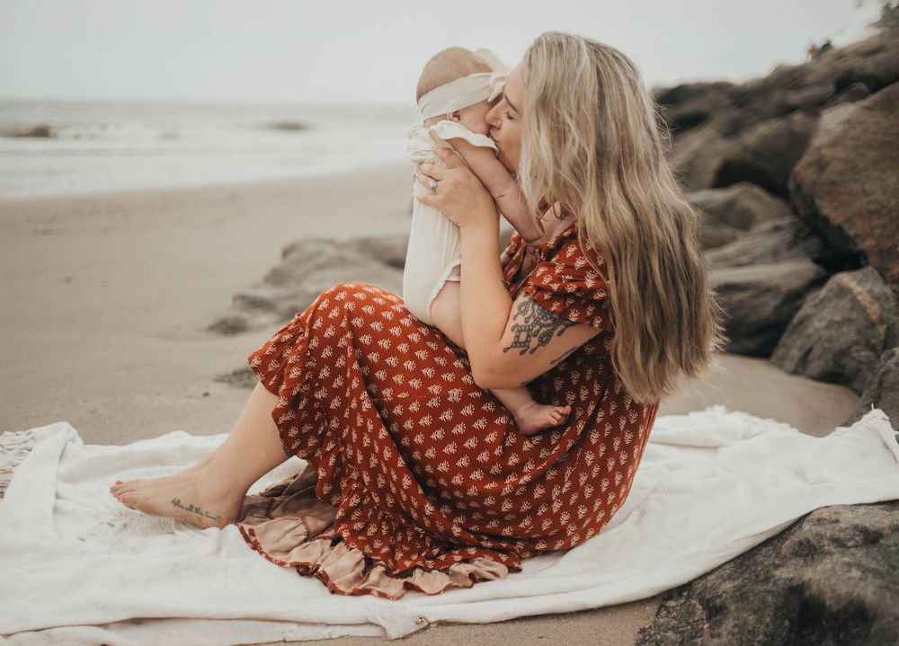 Mother in red floral dress kissing newborn baby on beach blanket with rocks and ocean in background.