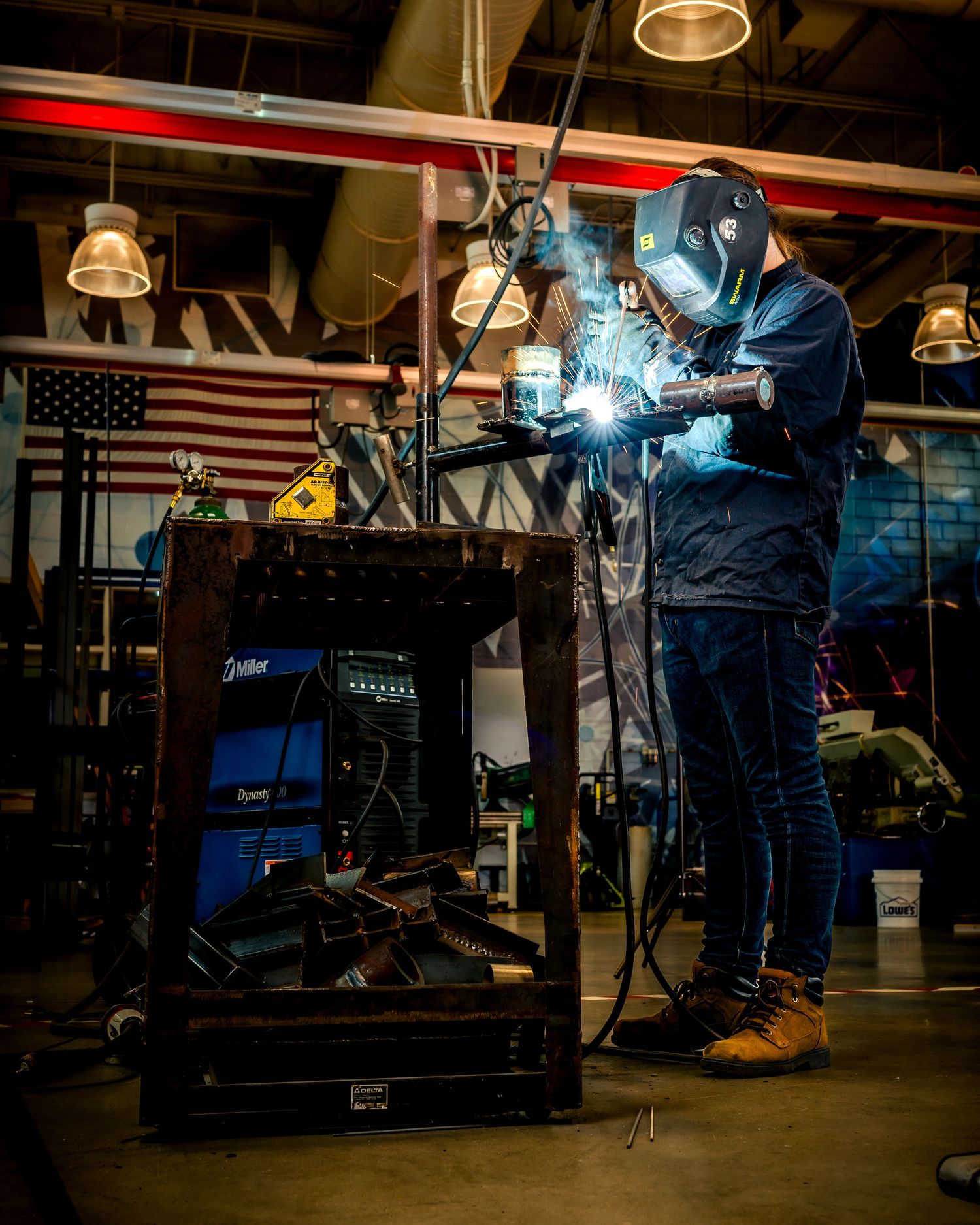 A welder in protective mask and gear creates bright sparks while working in an industrial workshop with an American flag backdrop.