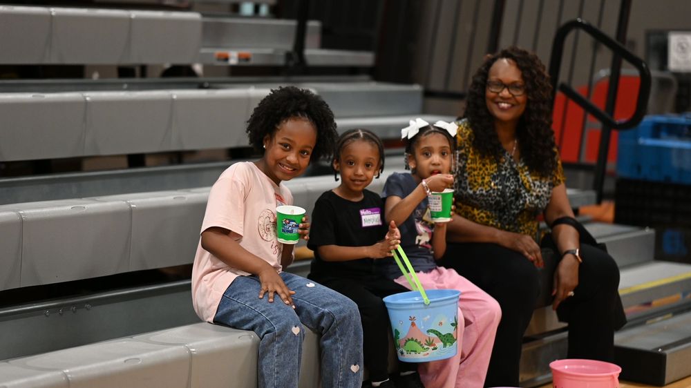 A woman and three young girls sit on bleachers smiling, holding green drinks at an indoor event.