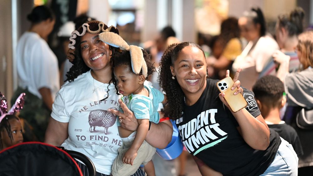 Two smiling women pose with a baby at a busy indoor event, one wearing a Student Dummy shirt.
