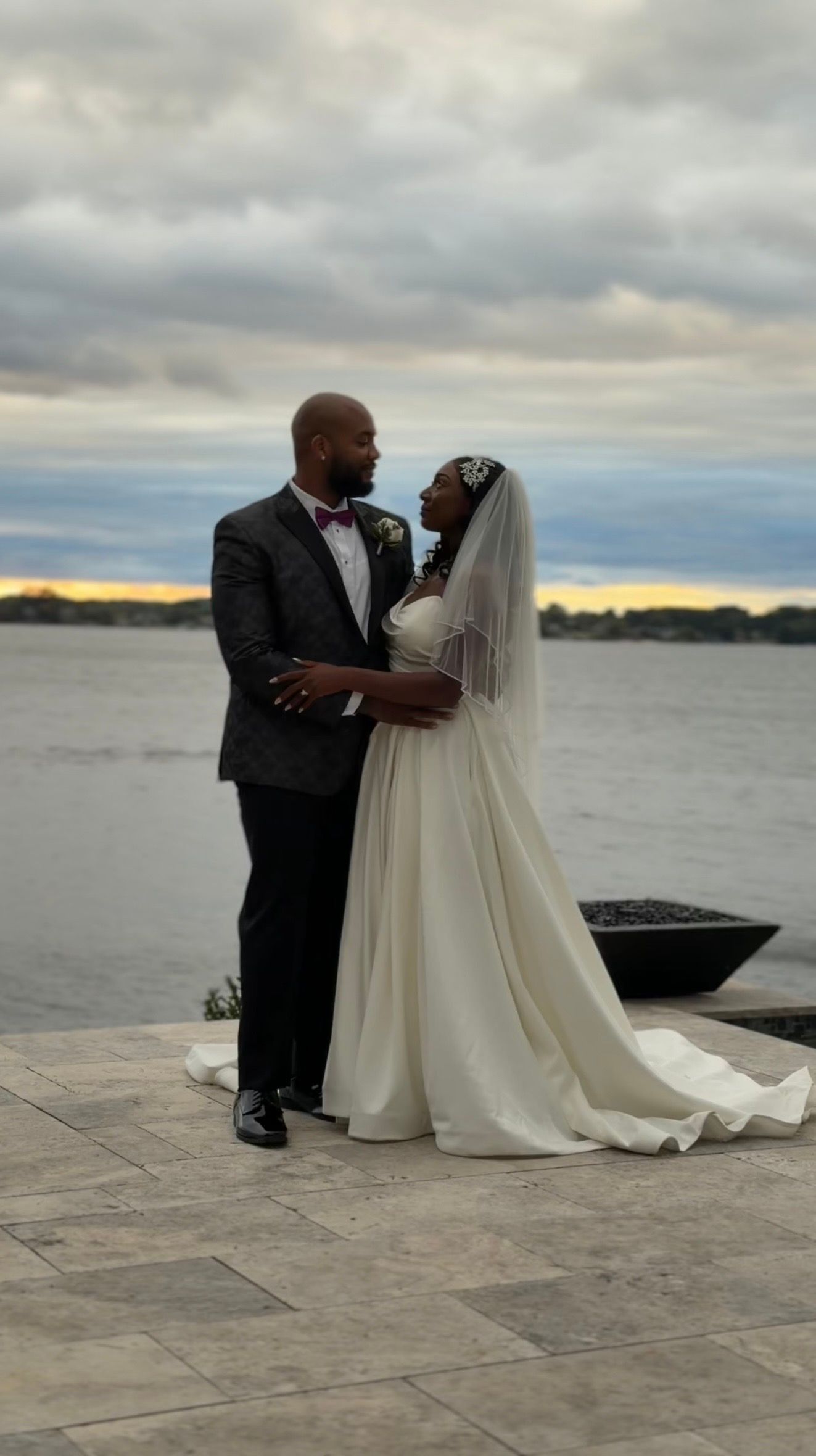 Bride in white gown and groom in gray suit share a romantic moment by the waterfront at sunset.