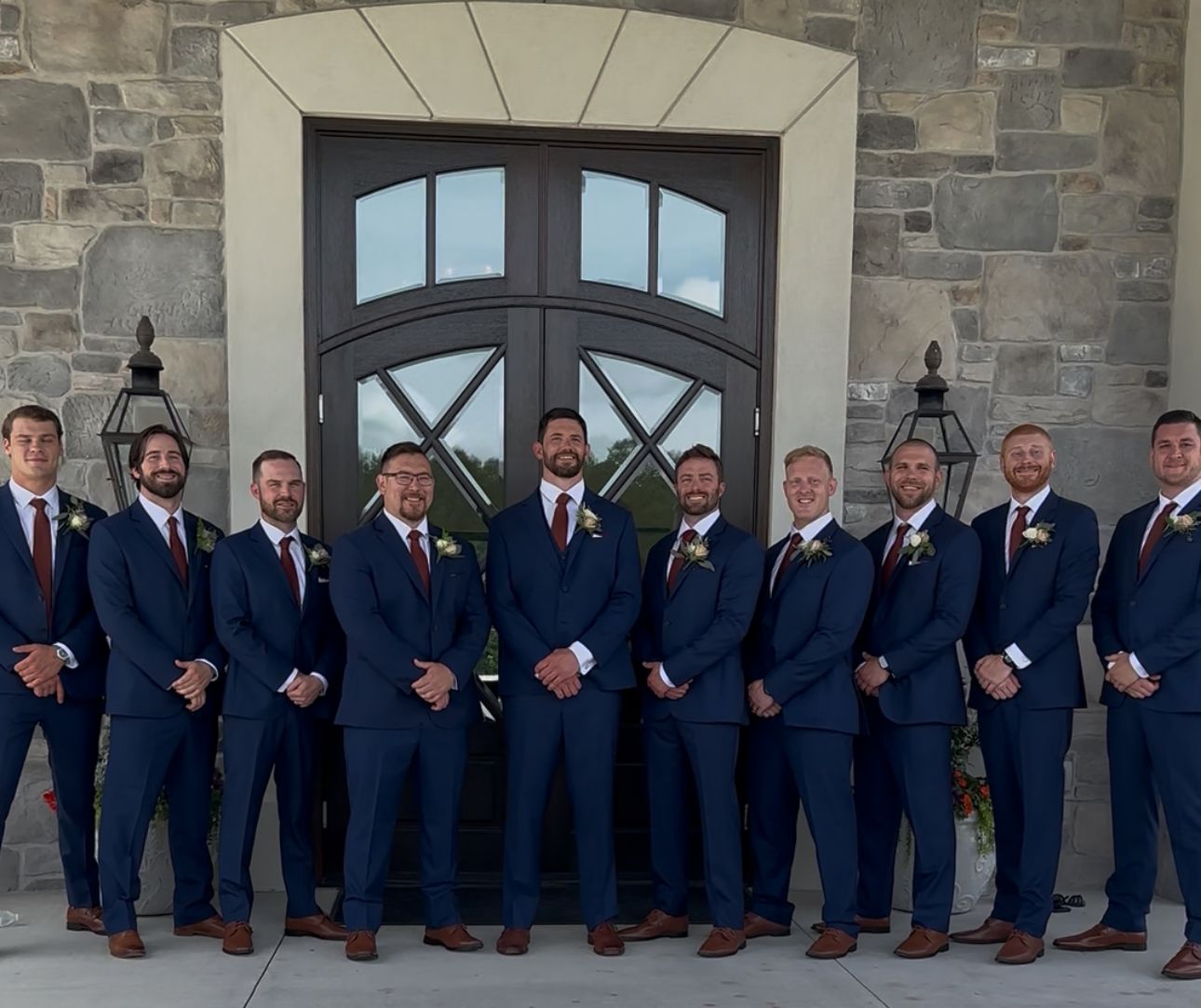 Ten groomsmen in navy suits with burgundy ties pose in front of stone building double doors at a wedding.