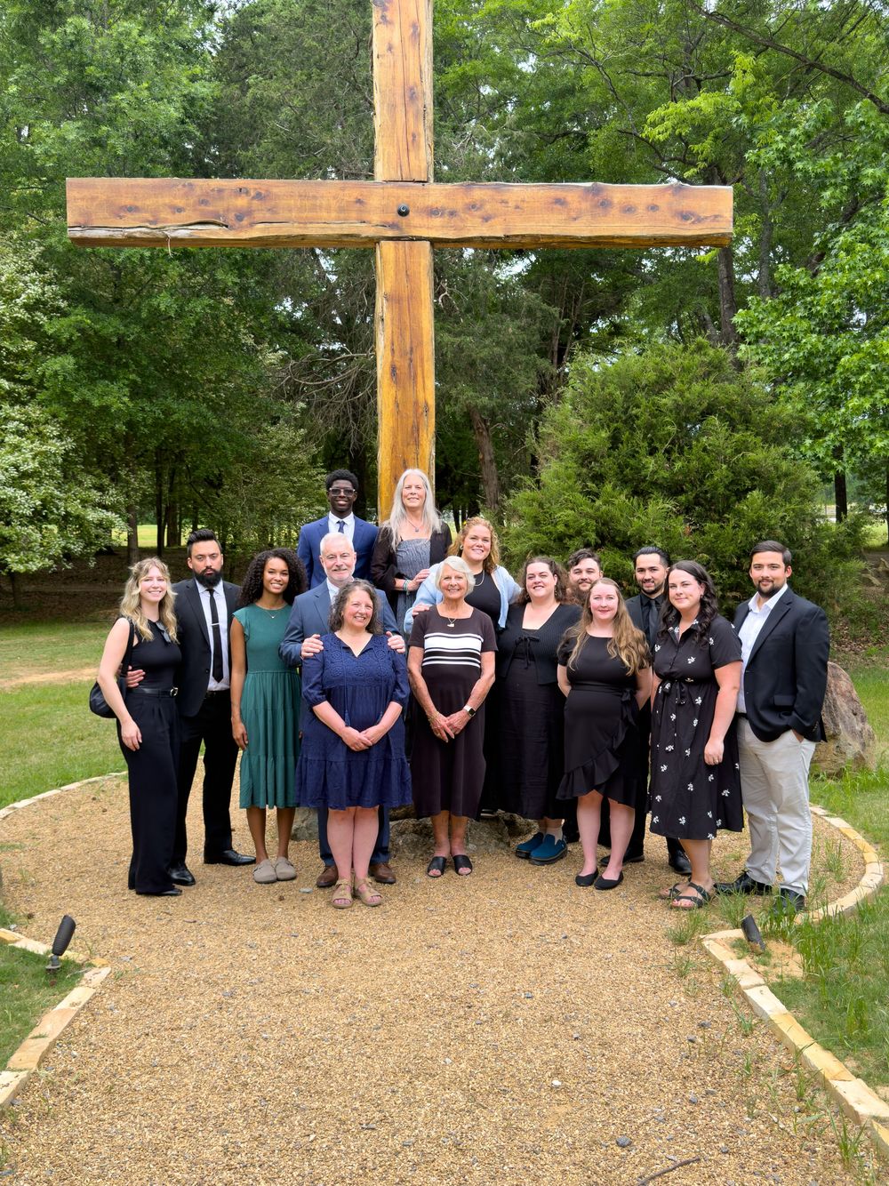 Group of formally dressed people posing together outdoors in front of a large wooden cross surrounded by trees.