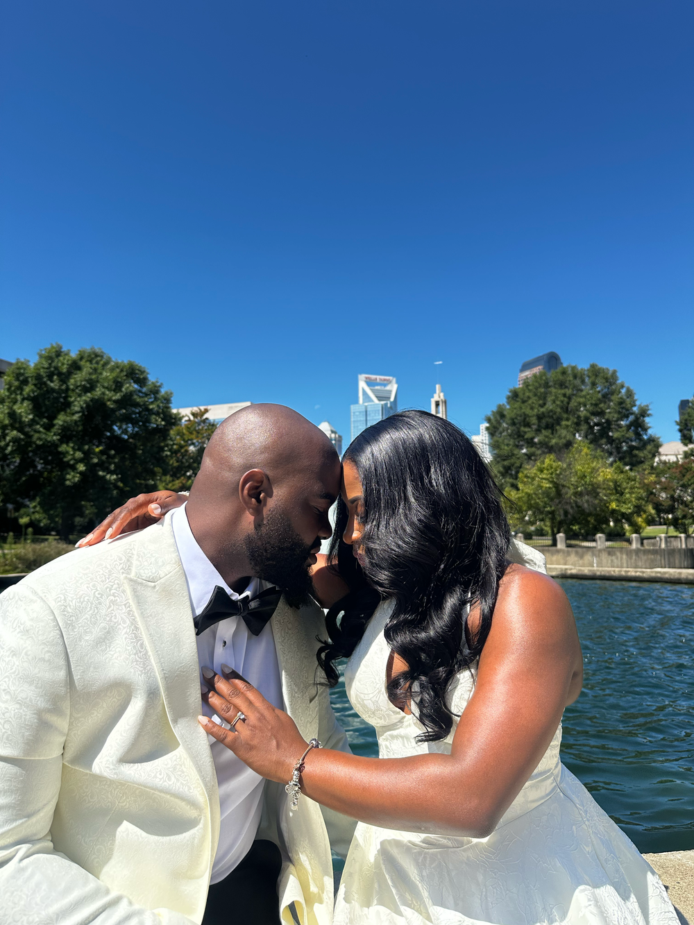 A couple in wedding attire shares a romantic moment by a city waterfront under a clear blue sky.