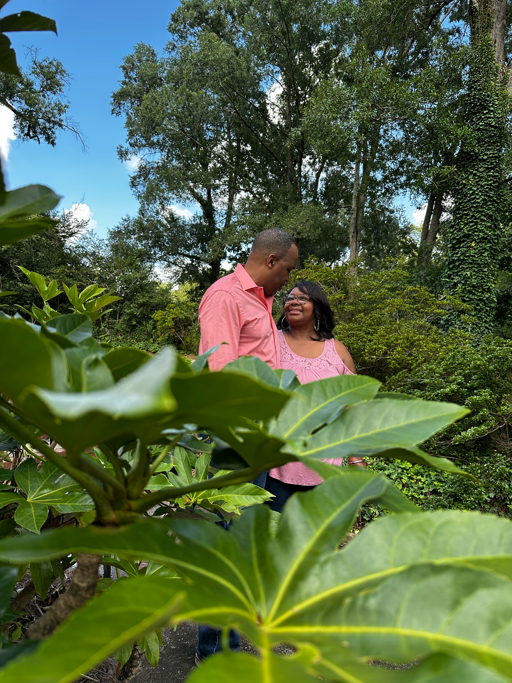 A couple in matching pink outfits share a romantic moment surrounded by lush green tropical plants and trees.