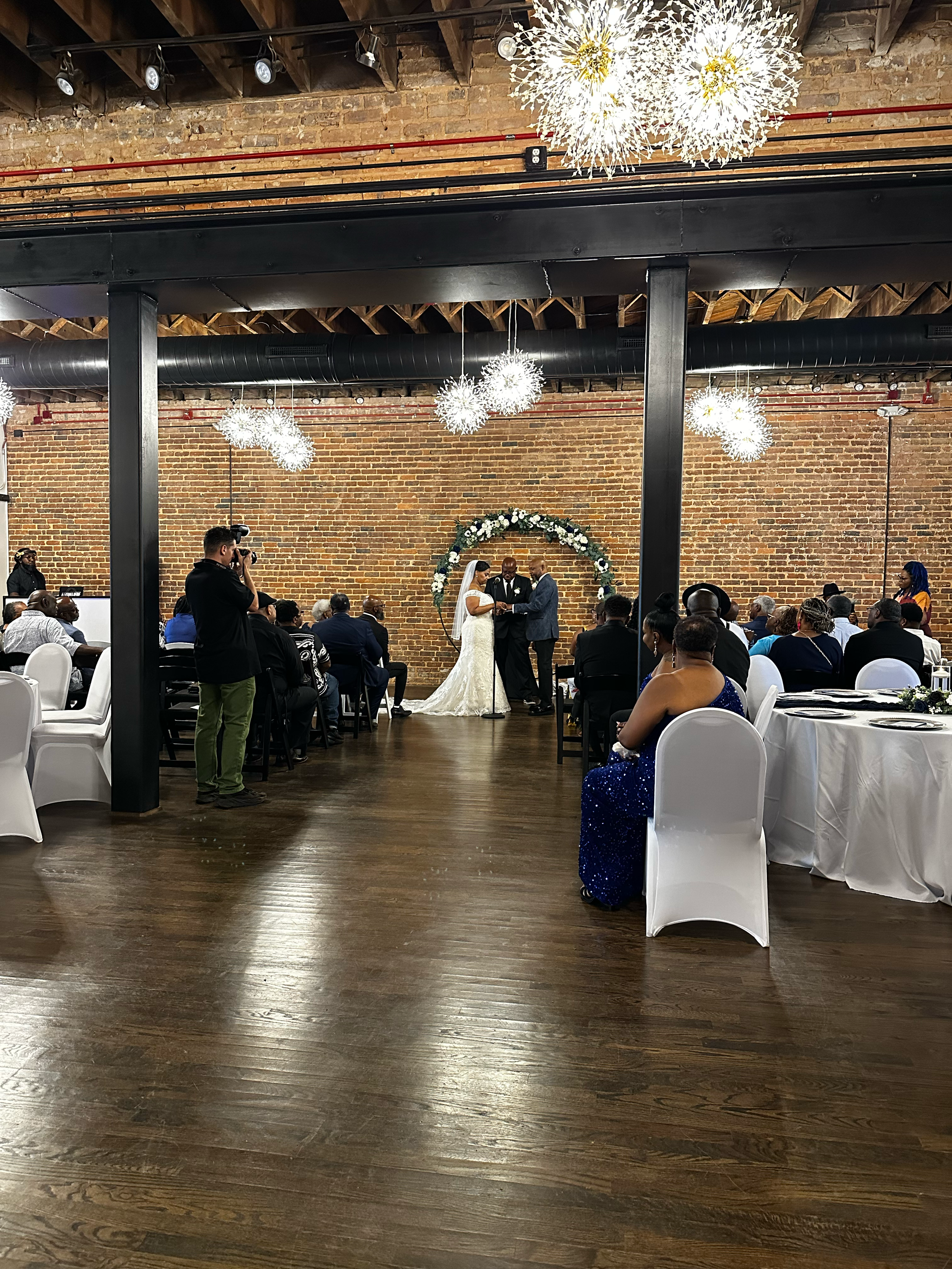 Wedding ceremony in rustic brick venue with chandelier lighting, guests seated at white-draped tables as couple exchanges vows.