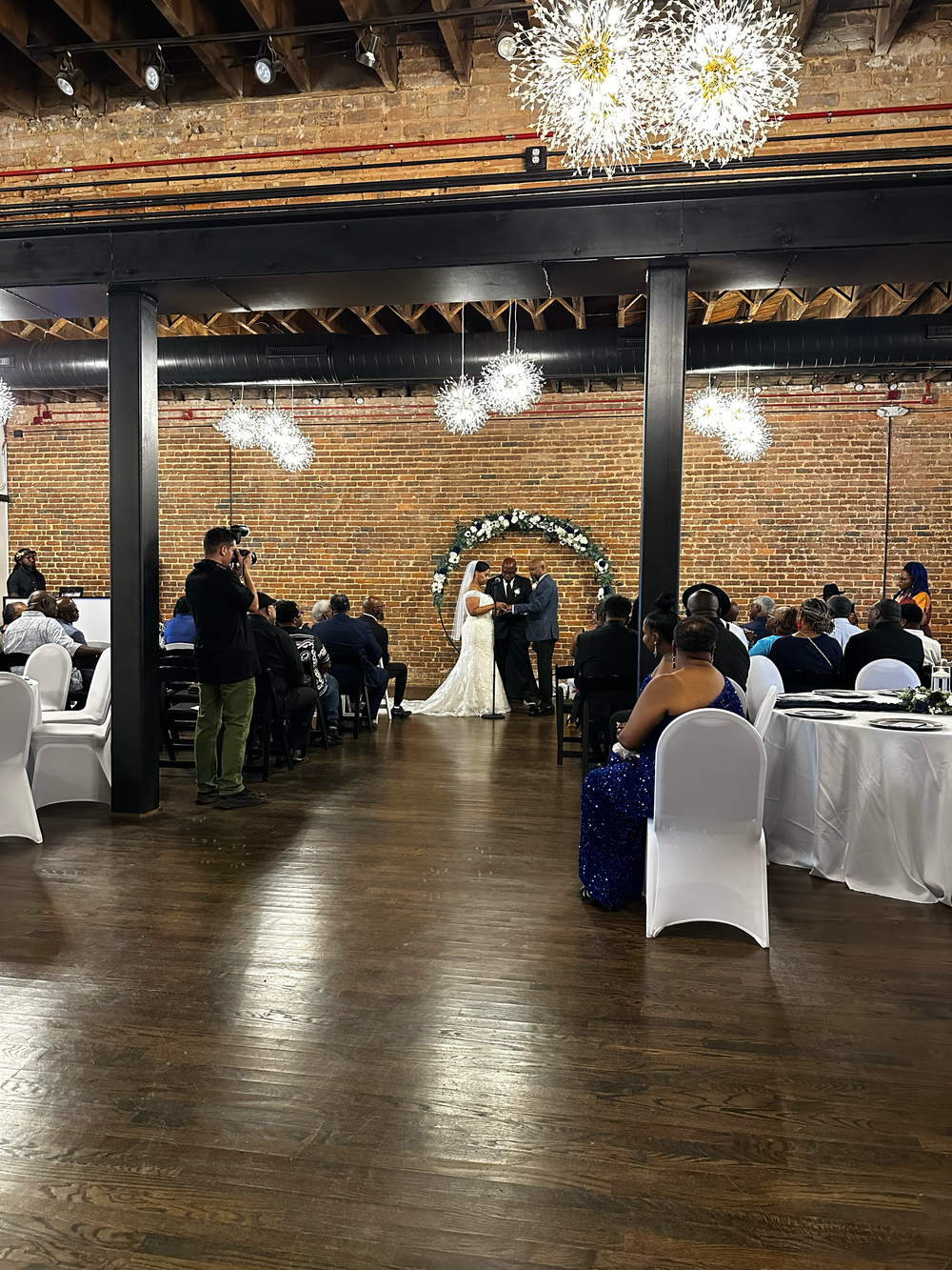 Wedding ceremony in rustic brick venue with chandelier lighting, guests seated at white-draped tables as couple exchanges vows.