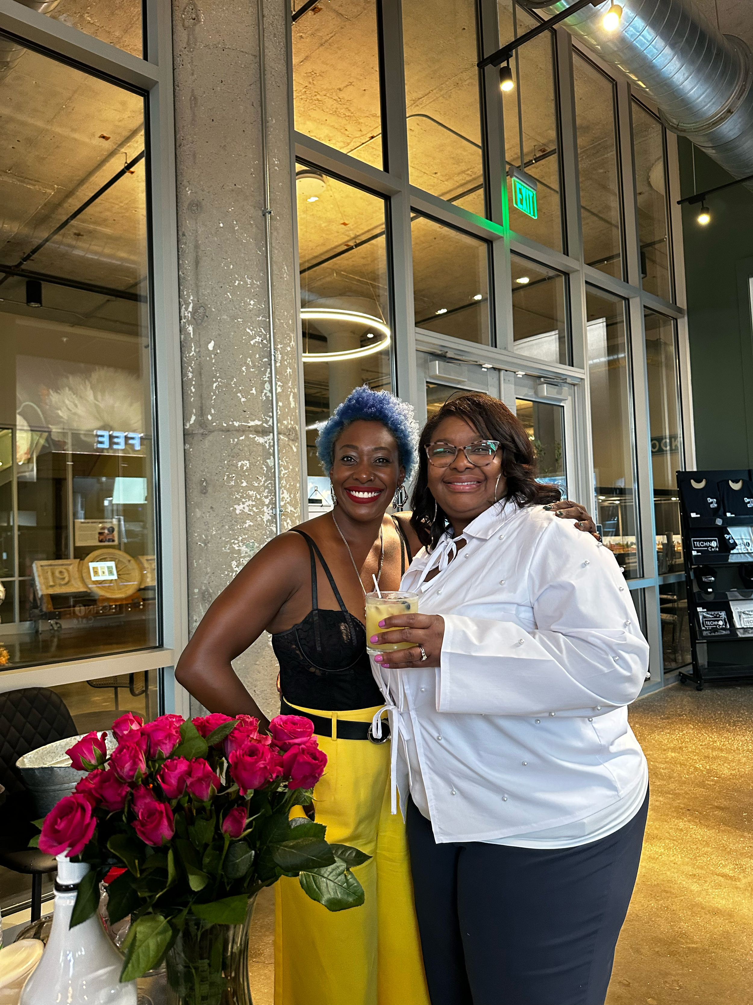 Two women smiling at an indoor event, one in a yellow skirt with blue hair, beside pink roses.