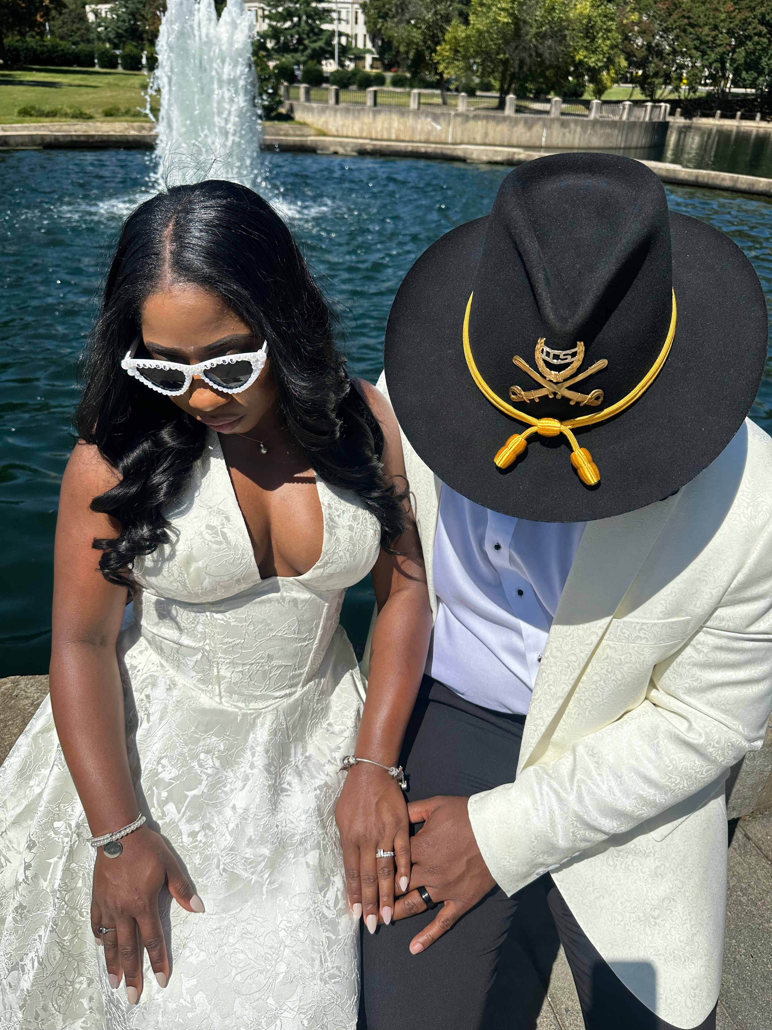 Couple in wedding attire poses by fountain, bride in white gown with sunglasses, groom in cream suit with black cowboy hat.