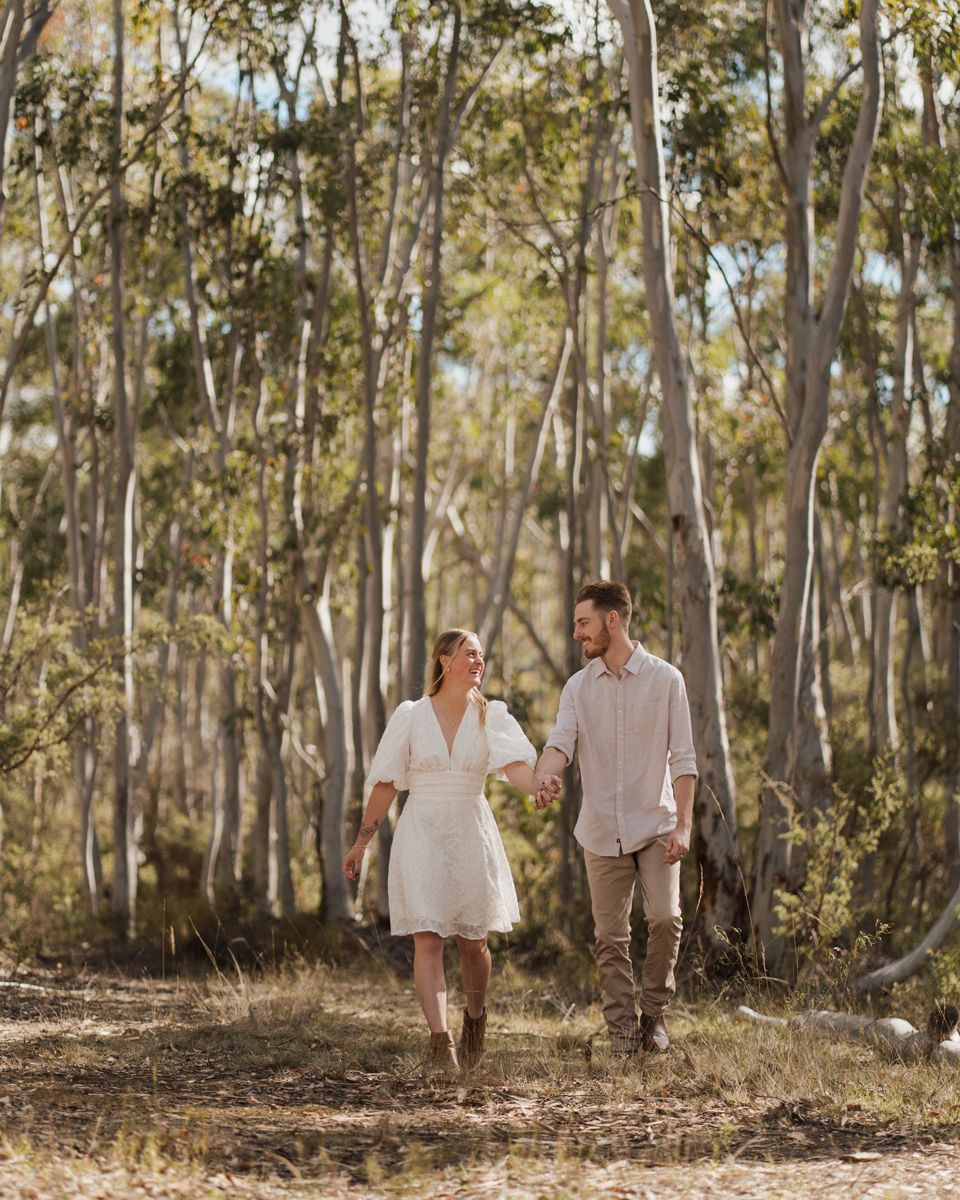 Couple holding hands walking through a sunlit eucalyptus forest, woman in white dress and man in casual attire in rouse hill photoshoot