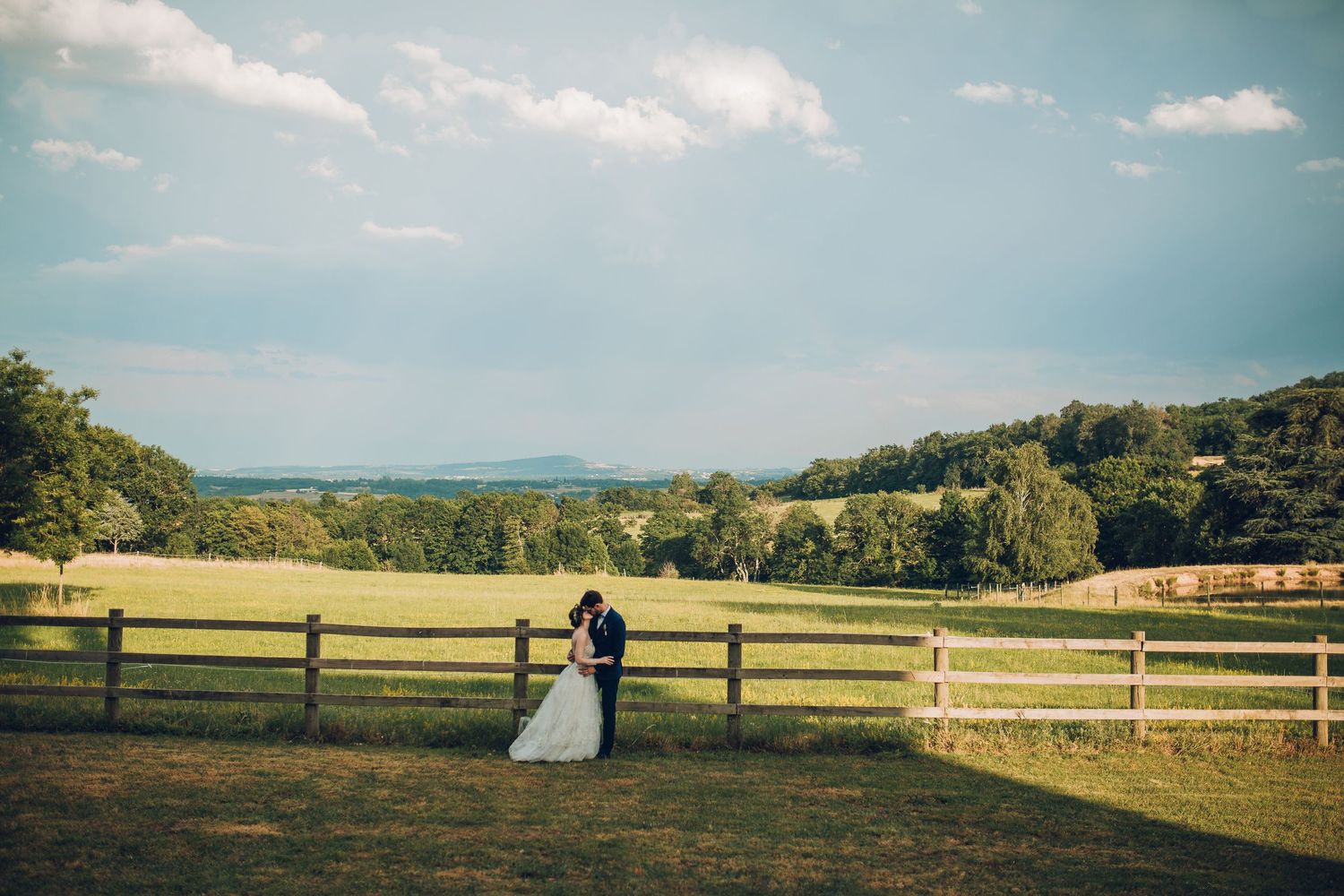 Bride and groom embracing by wooden fence in scenic countryside landscape.