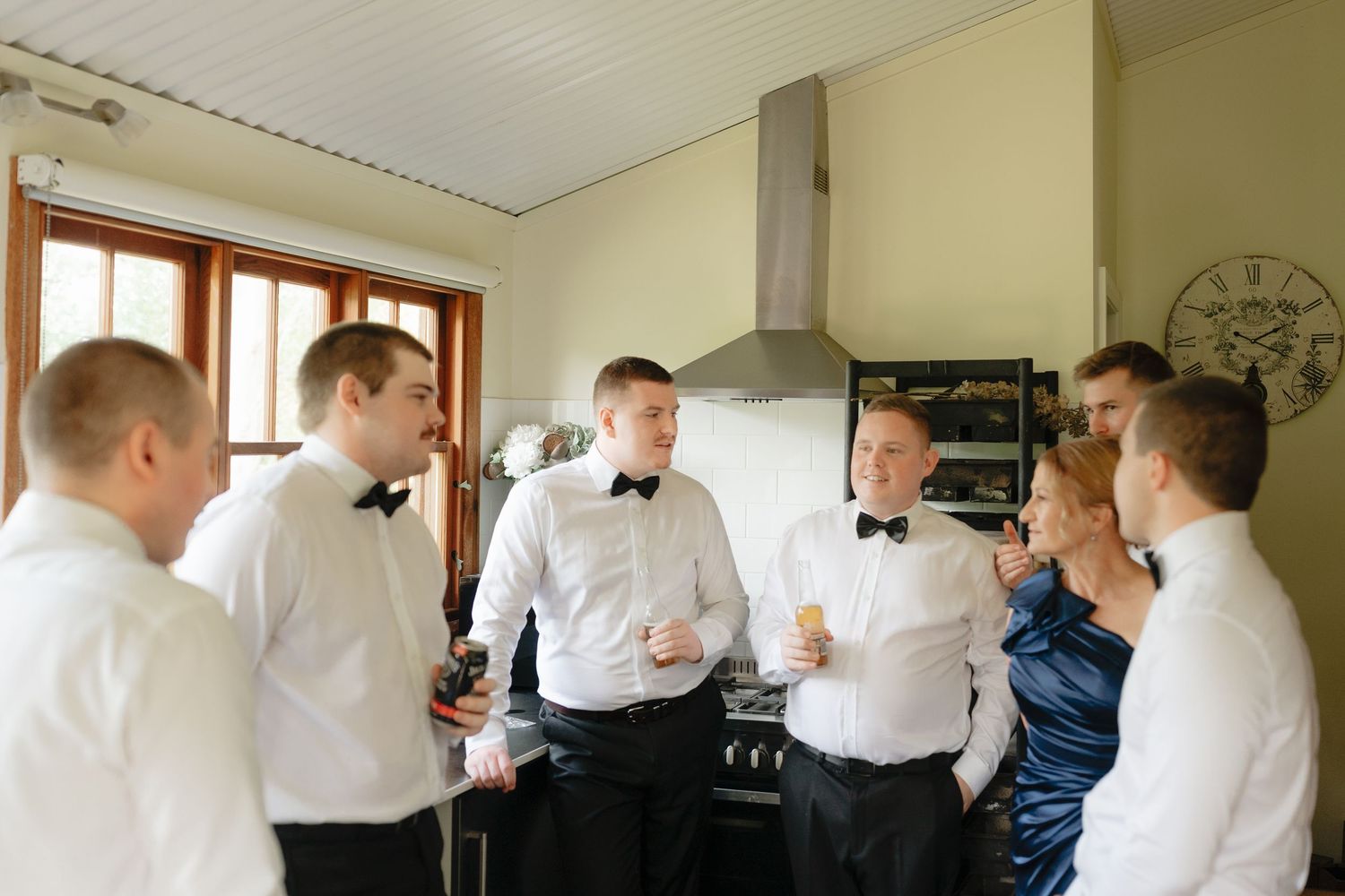 Groomsmen in white shirts and black bow ties socializing in a kitchen before a wedding ceremony.