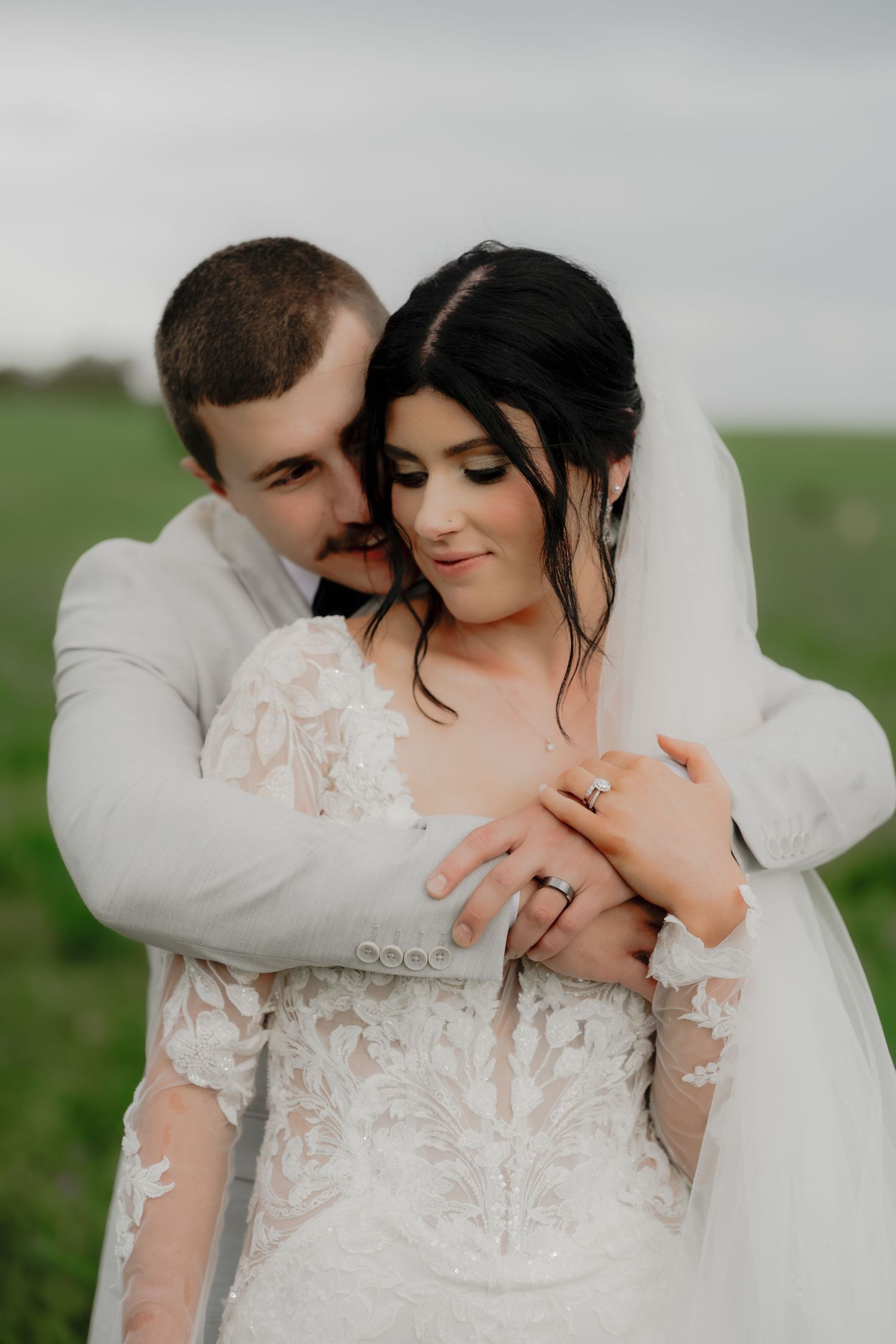 A groom in a gray suit embraces his bride in a lace wedding dress and veil outdoors on their wedding day.