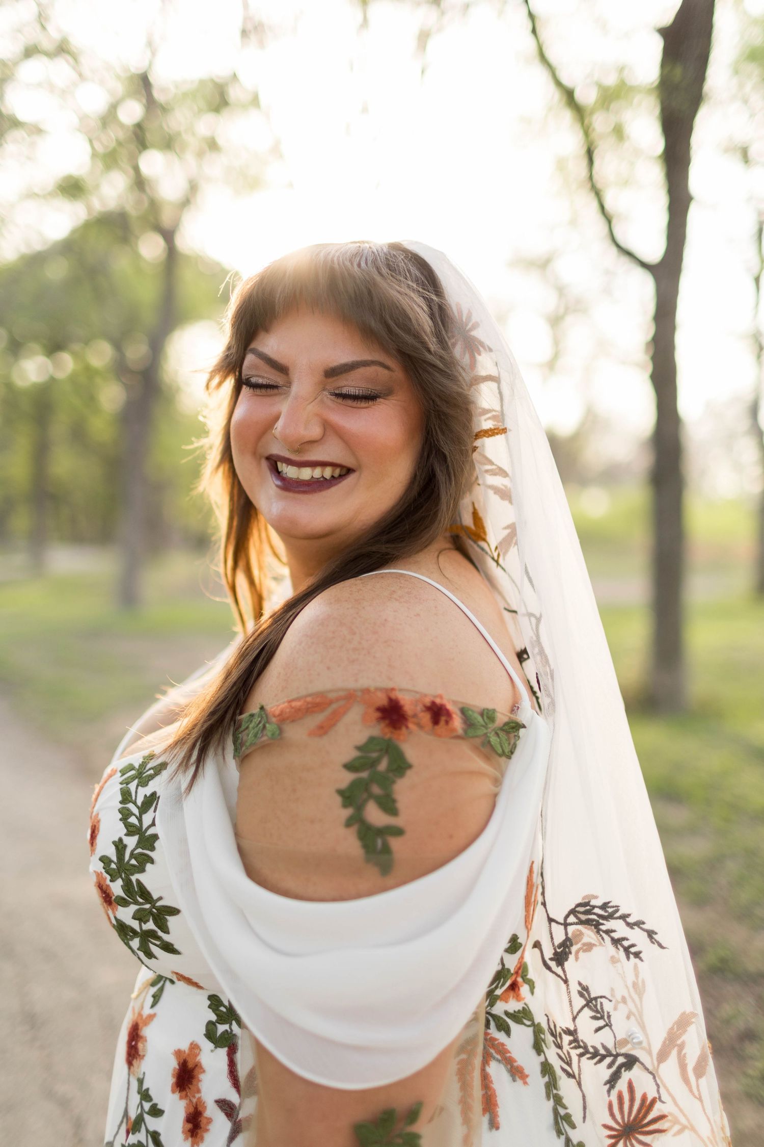 Smiling bride in floral embroidered wedding dress with veil, showcasing back tattoos outdoors in golden hour light.
