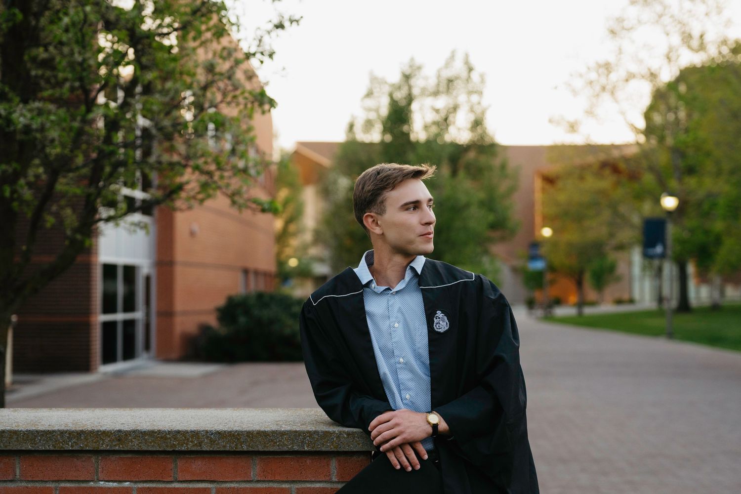 Young man in graduation gown standing on a college campus pathway surrounded by trees and brick buildings.