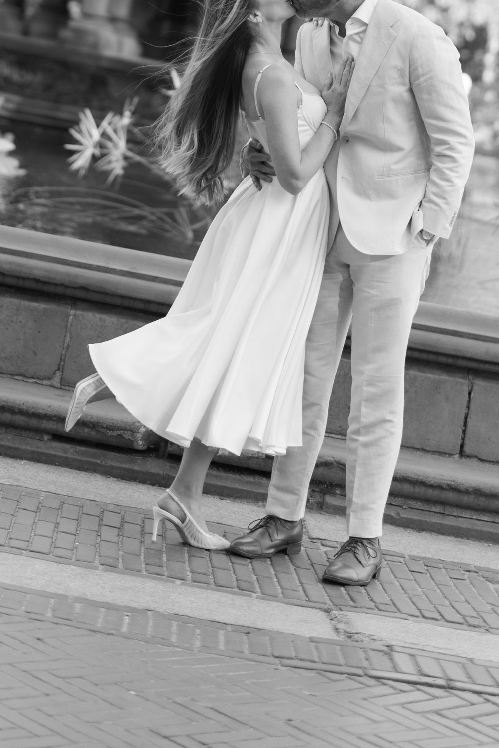 Black and white photo of a couple kissing near a fountain, woman in white dress and man in light suit on brick pavement.