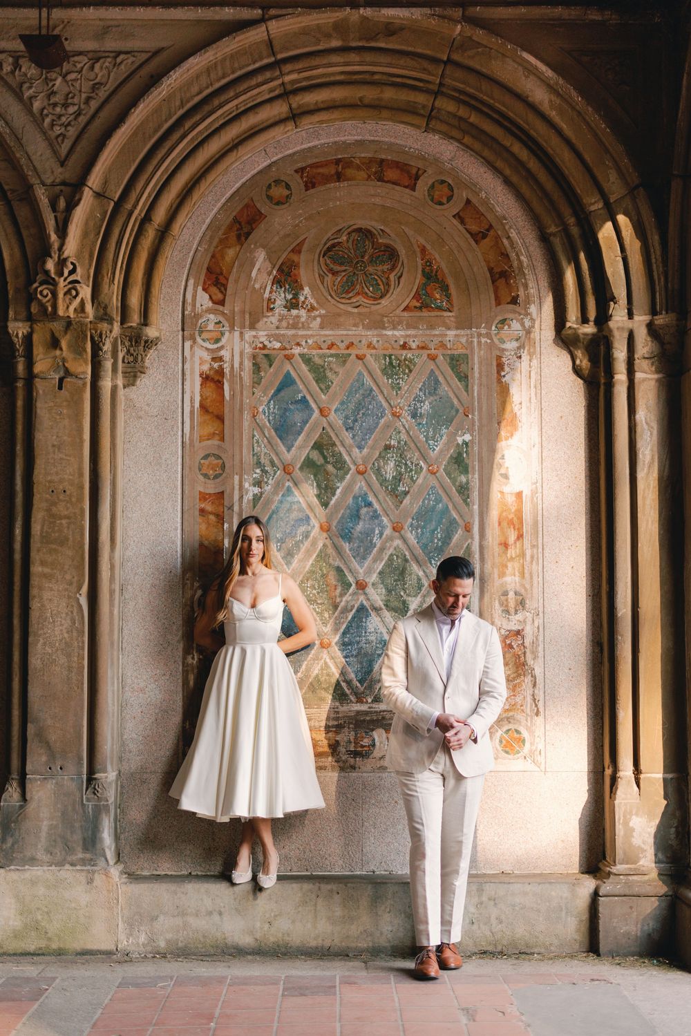 Bride and groom pose under ornate stone arch with colorful mosaic at Bethesda Terrace, Central Park NYC.