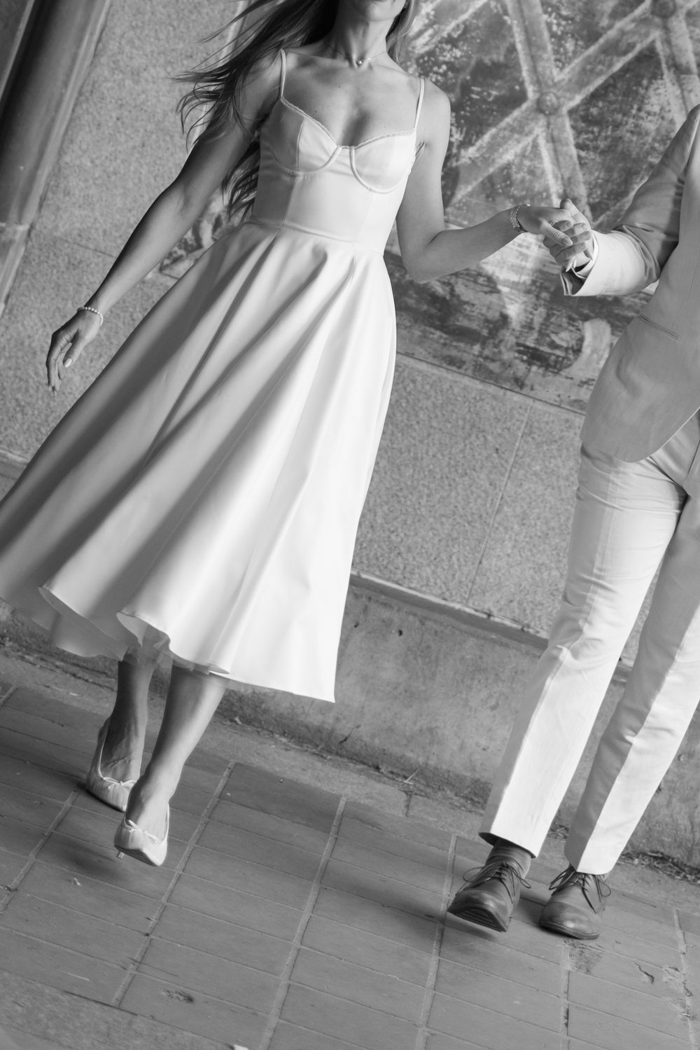 Black and white photo of a couple walking hand in hand, woman in white midi dress and man in light suit on cobblestone.
