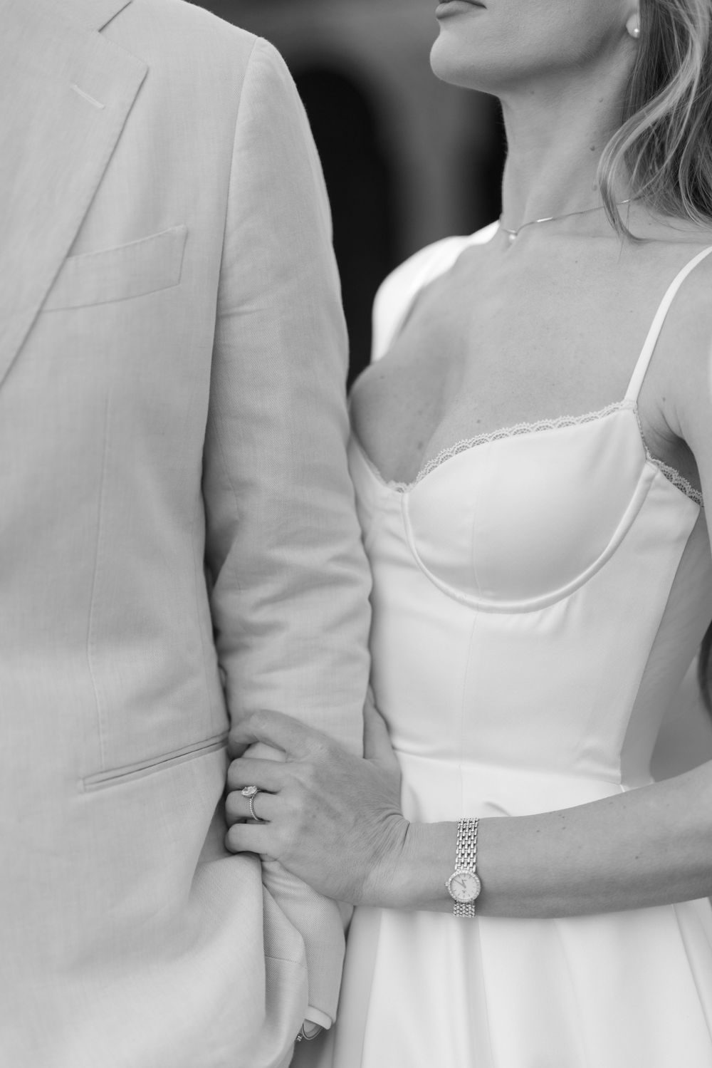 Black and white close-up of bride in white spaghetti strap gown holding grooms arm, wearing a watch.