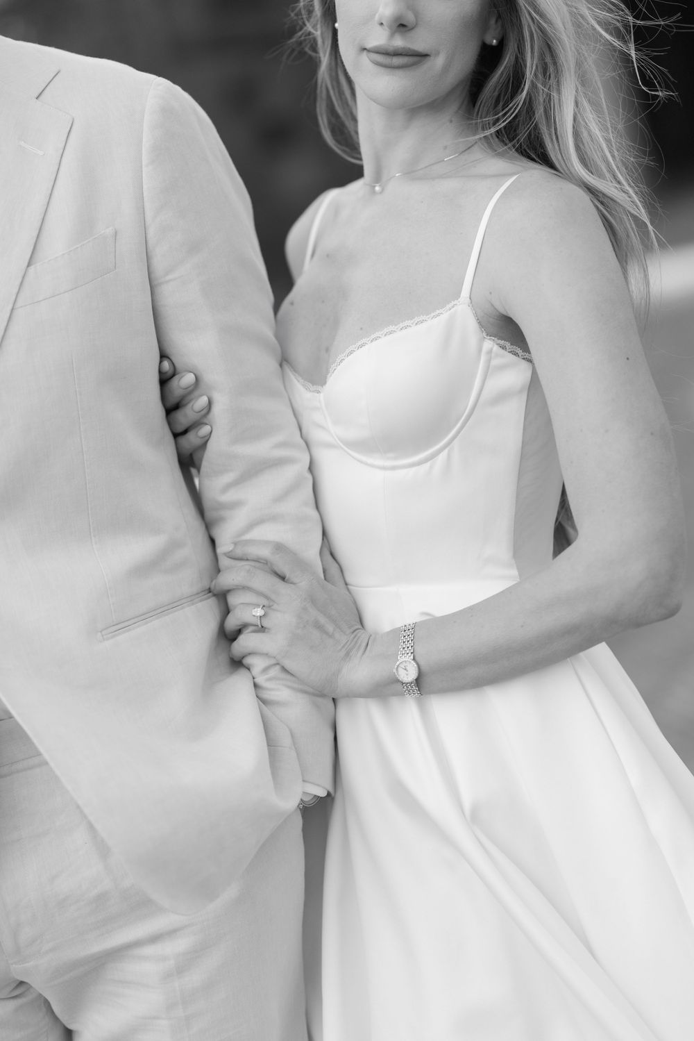Black and white close-up of bride in spaghetti strap gown holding grooms arm in beige suit.