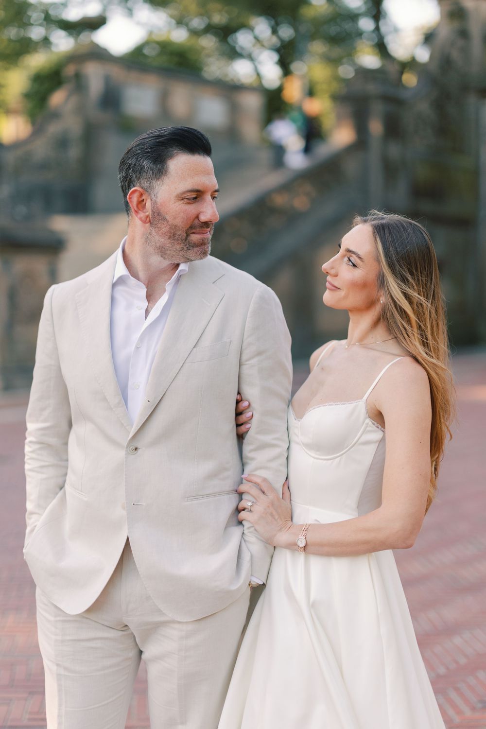 Stylish couple in matching cream outfits sharing a loving glance during an engagement shoot at Central Park's Bethesda Terrace.