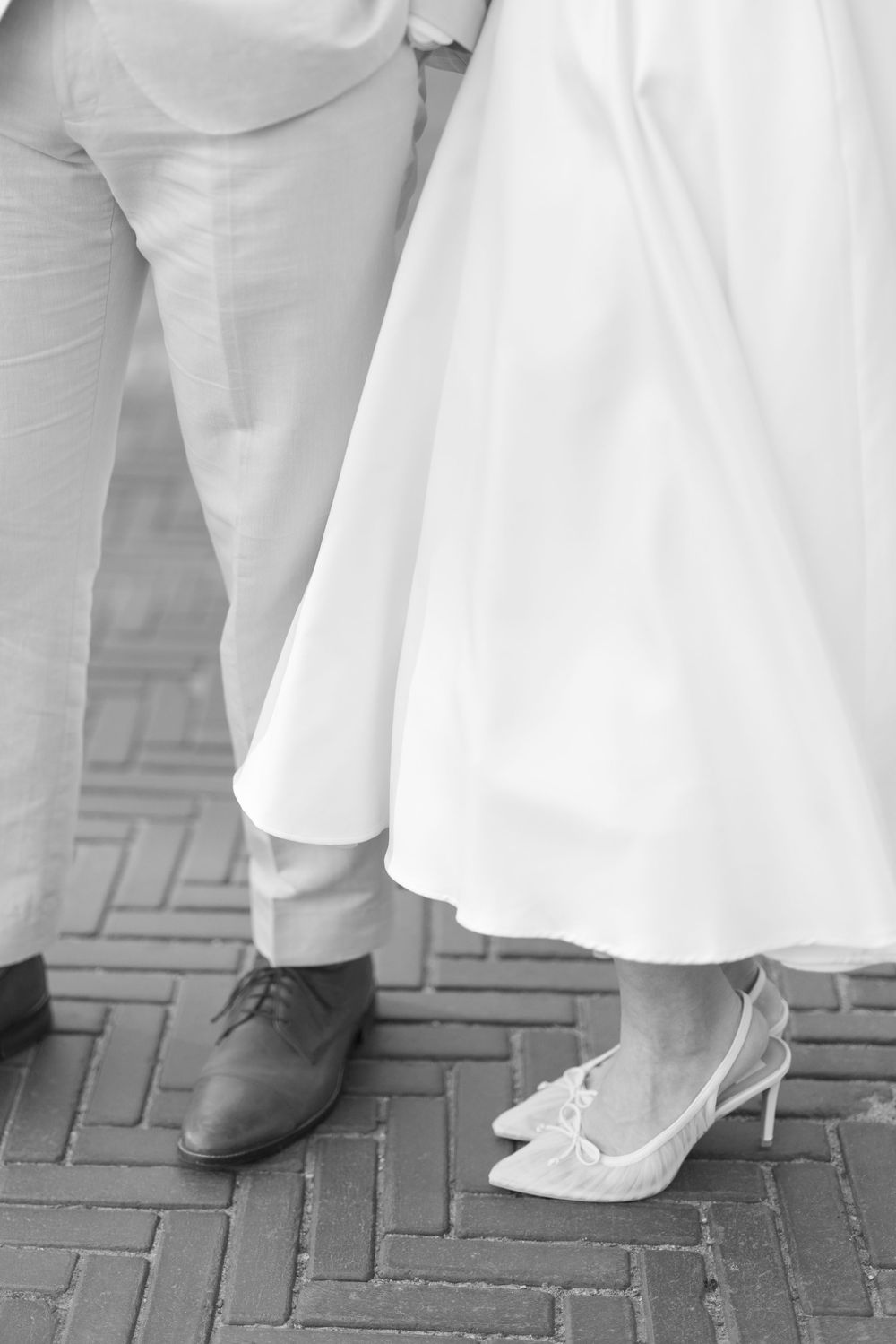 Black and white photo of bride in white tea-length dress with lace heels standing beside groom in gray suit on brick pavement.