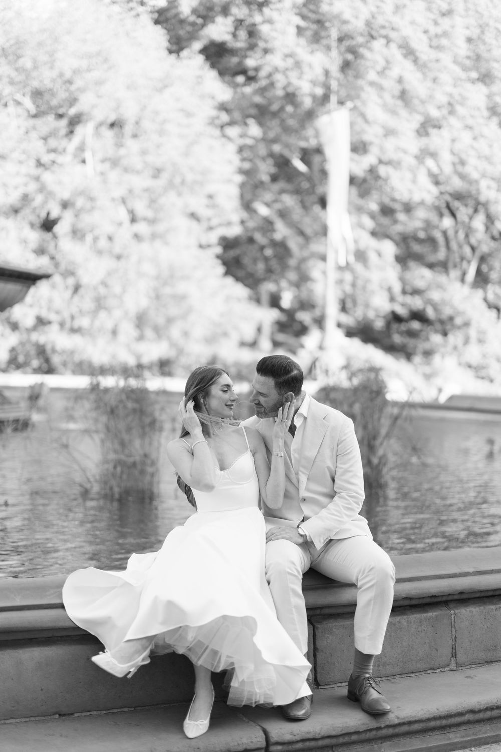 Black and white photo of bride and groom sitting by a Central Park fountain, sharing a romantic moment.